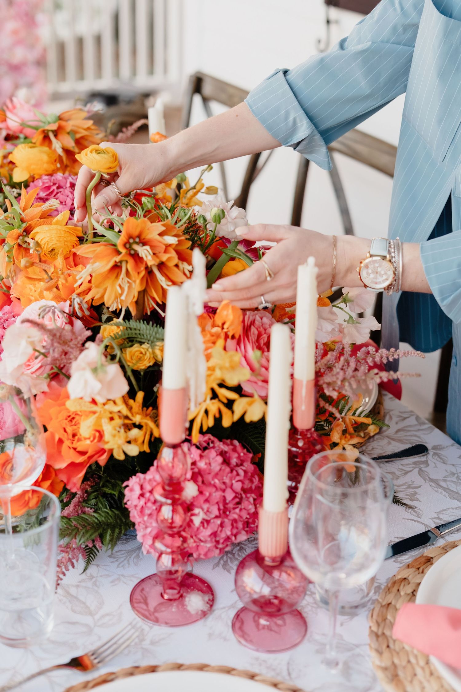 Hands arrange vibrant orange and pink flowers with blush candles on an elegant table setting with wine glasses.