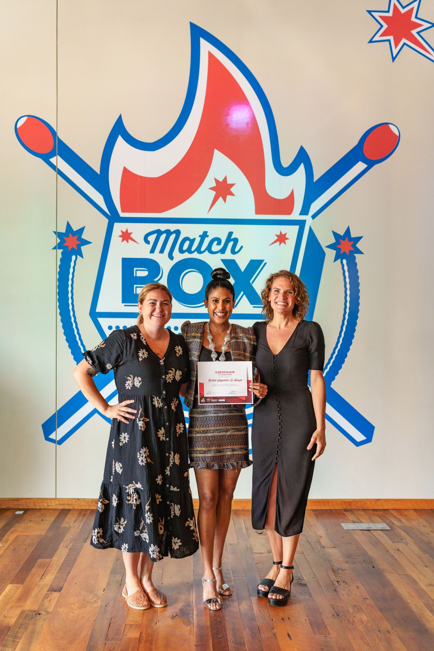 Three people stand posing together in front of a blue and red logo with crossed skis on a wooden floor.