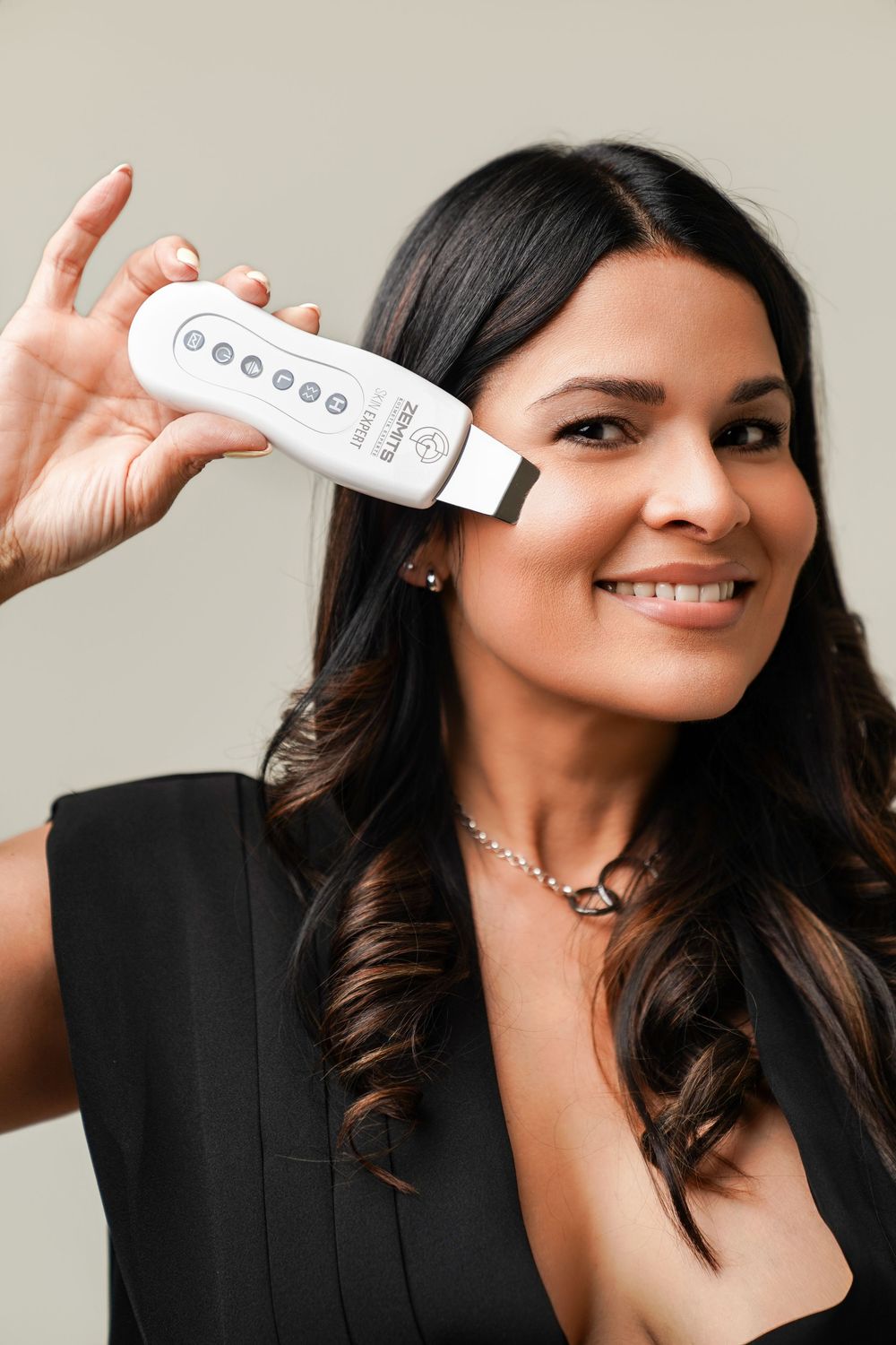 Smiling woman holding a white facial skincare device near her face, wearing a black top and pearl necklace.