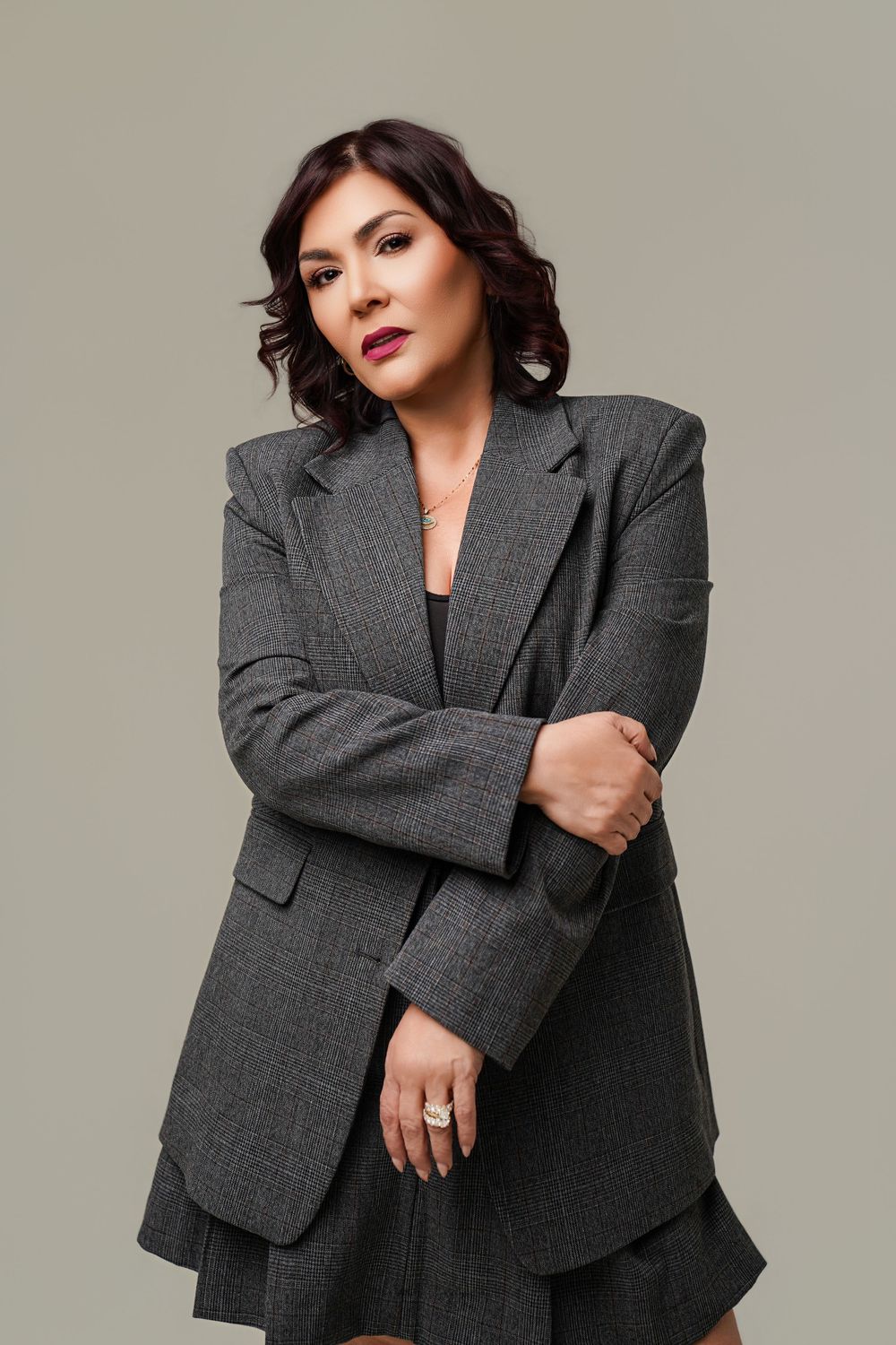 Woman in gray oversized blazer and skirt poses confidently against neutral background with bold red lips.