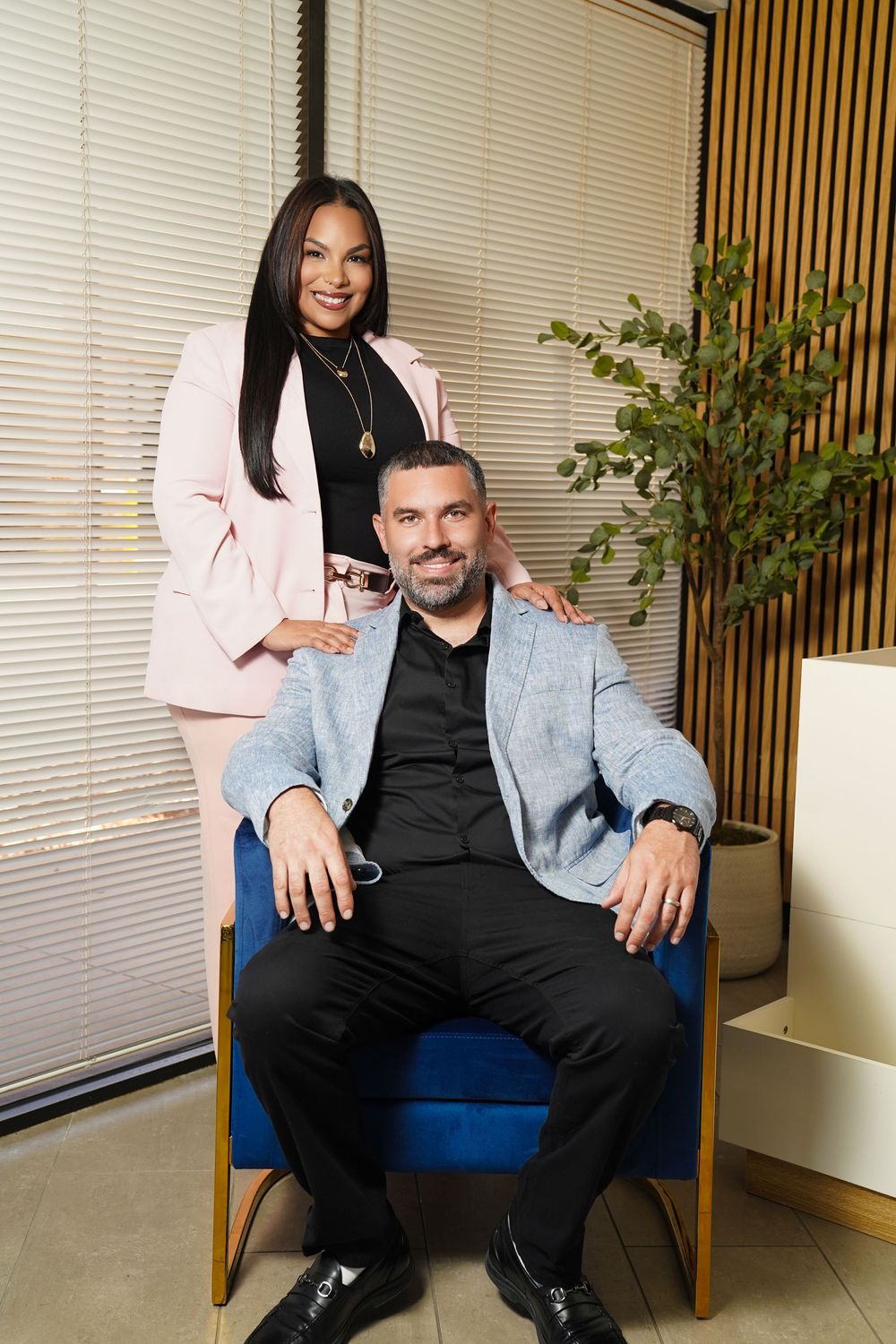 Professional man and woman posing in modern office, woman in pink blazer standing behind seated man in gray jacket.