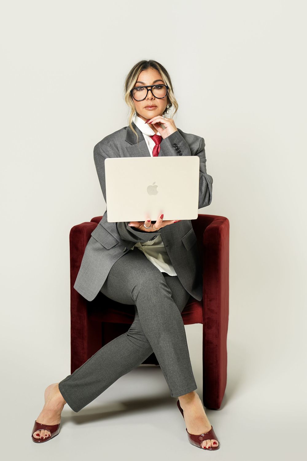 Woman in gray suit and red tie sits in burgundy chair with Apple laptop, wearing glasses, on light background.