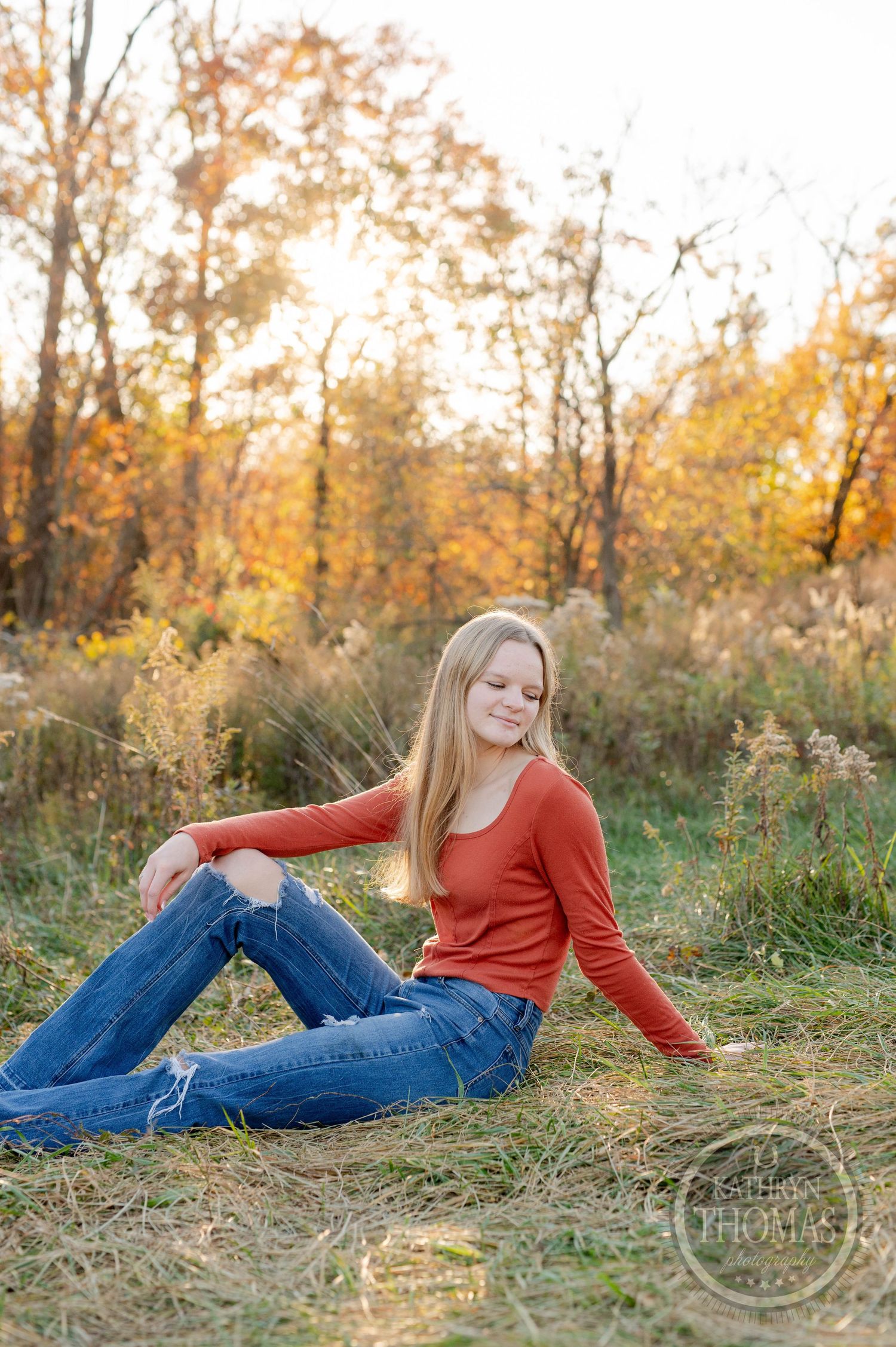 Relaxed autumn portrait sitting in golden grass wearing orange sweater and distressed denim.