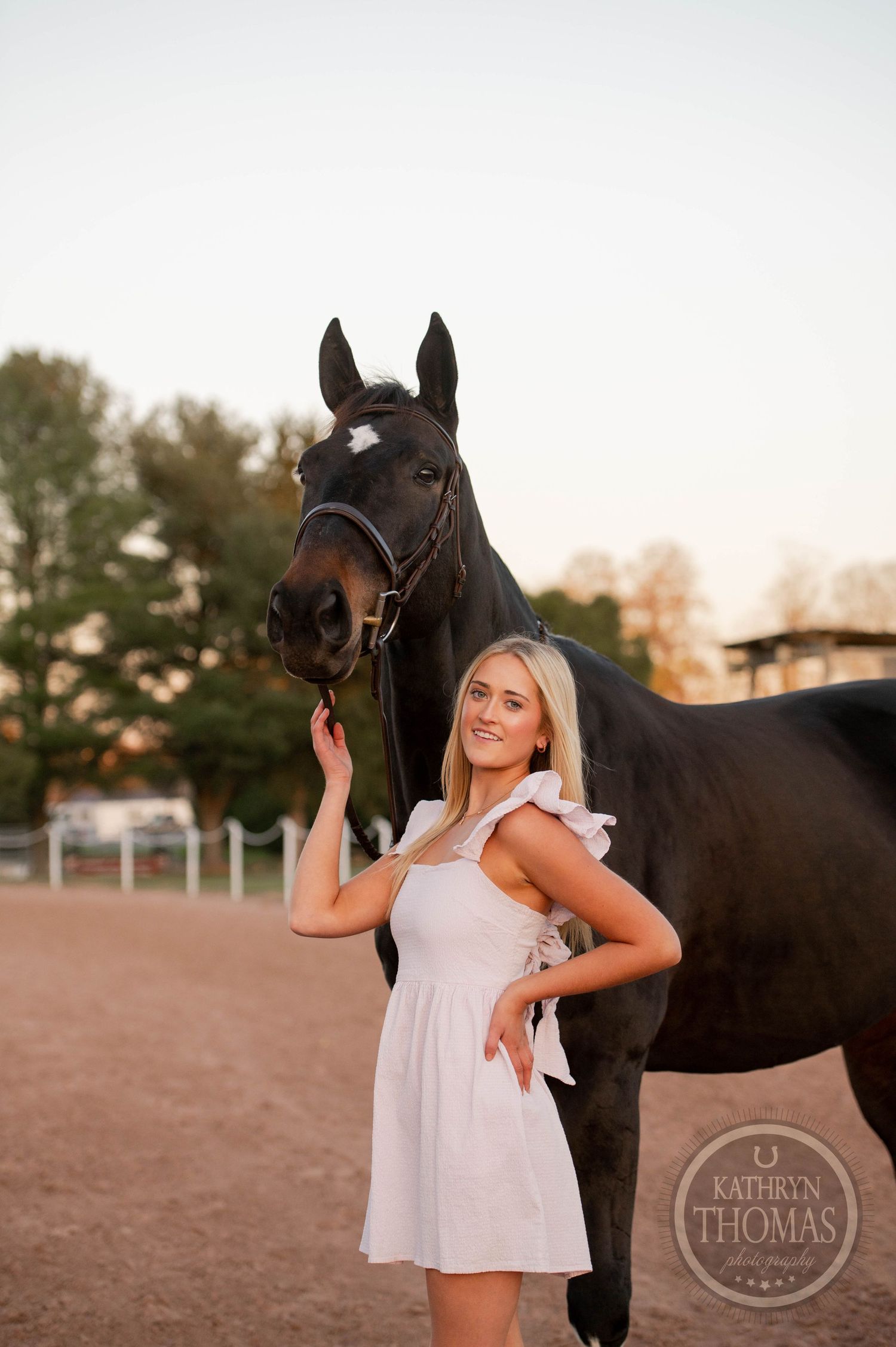A person in a white dress poses with a black horse during sunset.