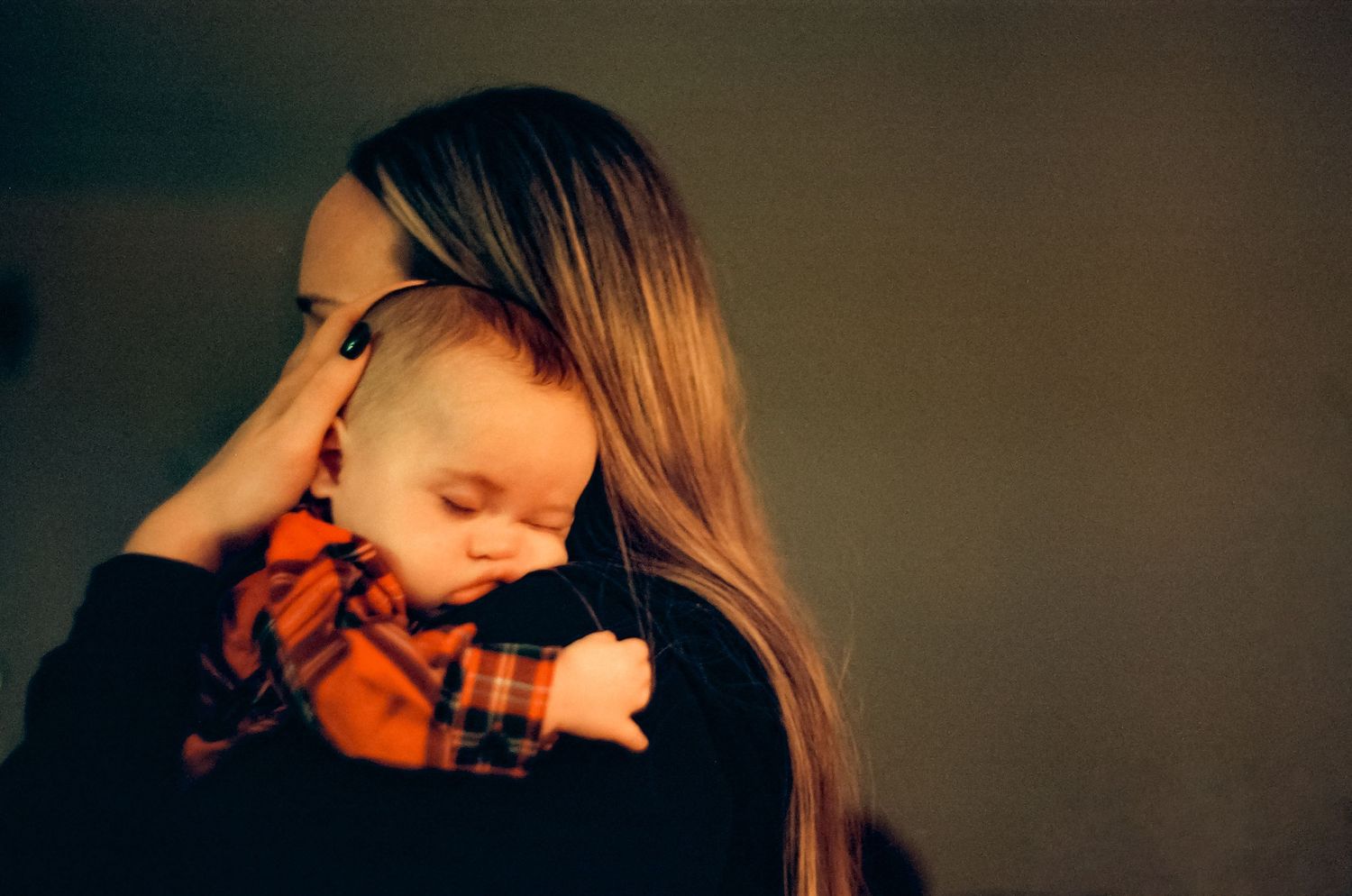 A sleeping baby in orange plaid rests on someone's shoulder, captured in dim lighting.