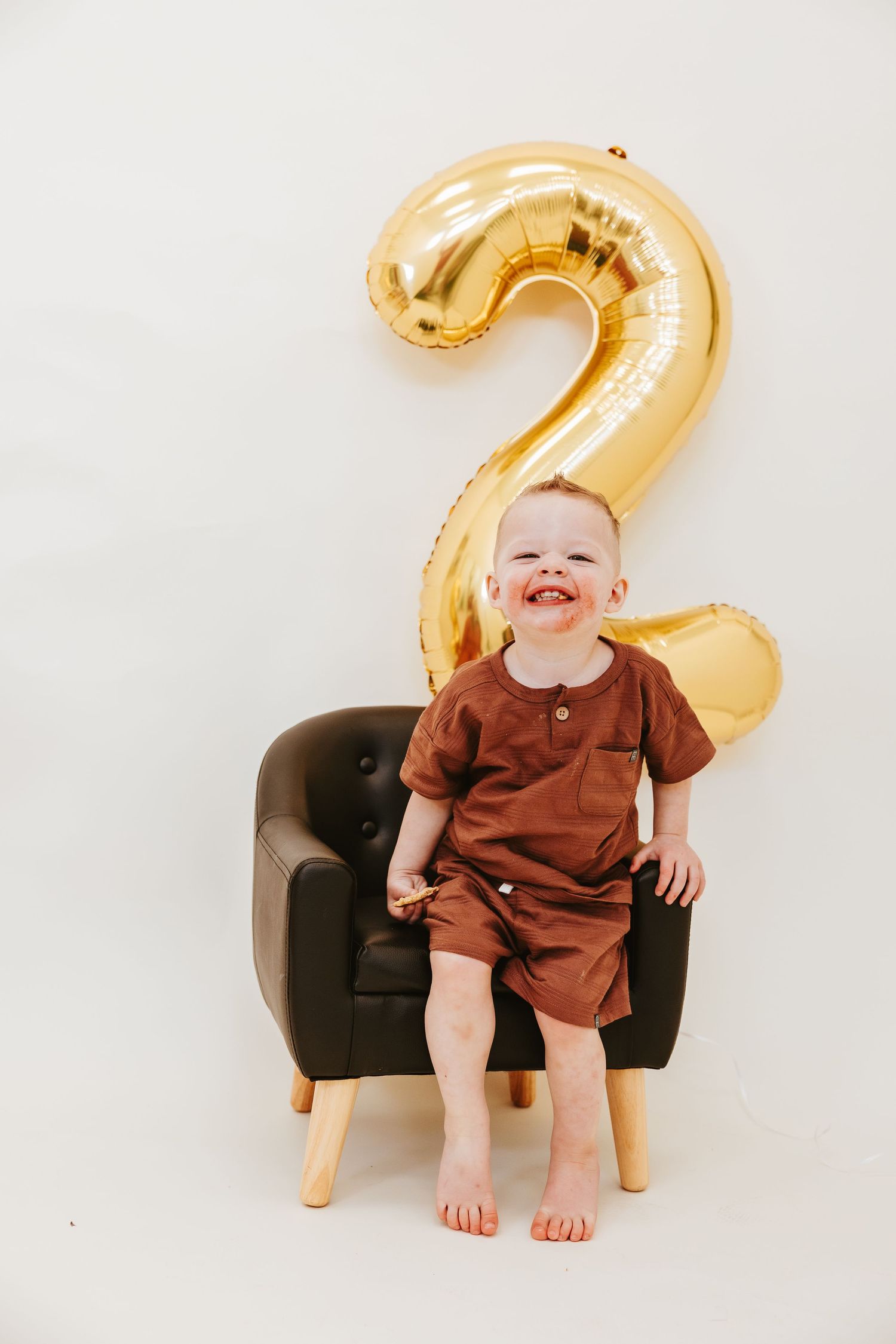 A joyful toddler in brown clothing sits on a dark chair with a gold number 2 balloon behind.