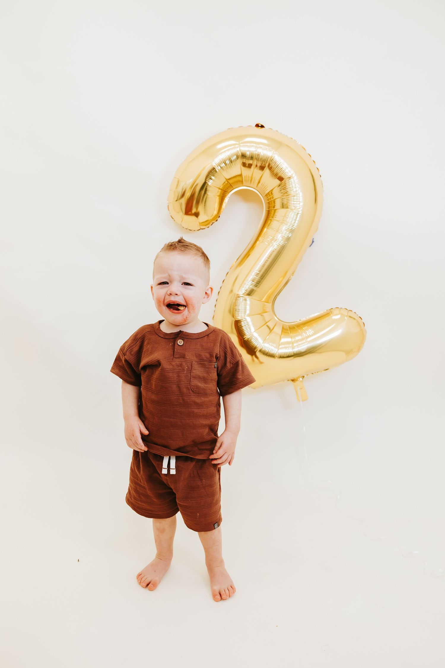 A young toddler in brown clothing stands happily next to a large gold number 2 balloon against a white background.