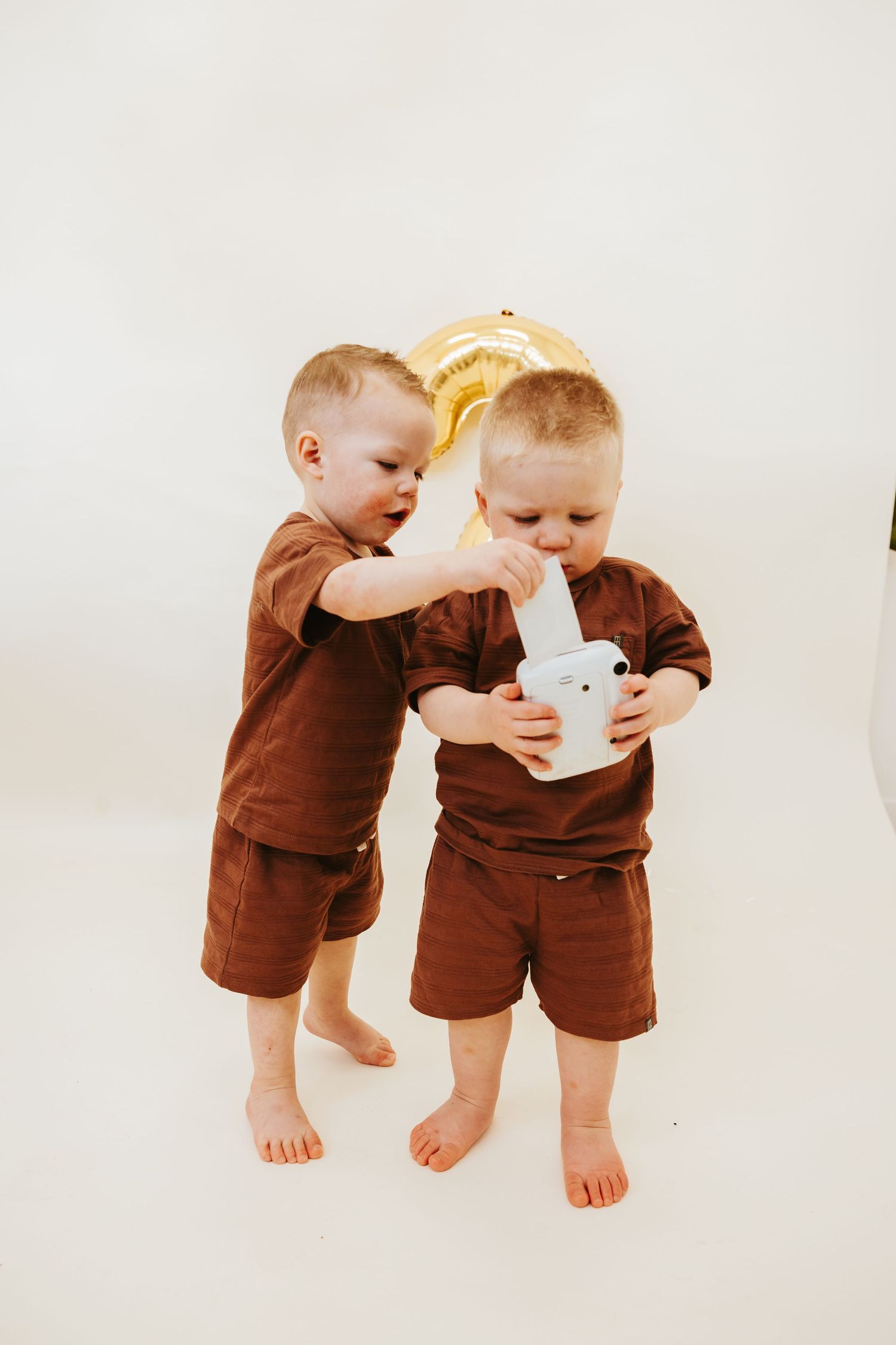 Two toddlers in matching brown outfits playing with a white toy against a white background.