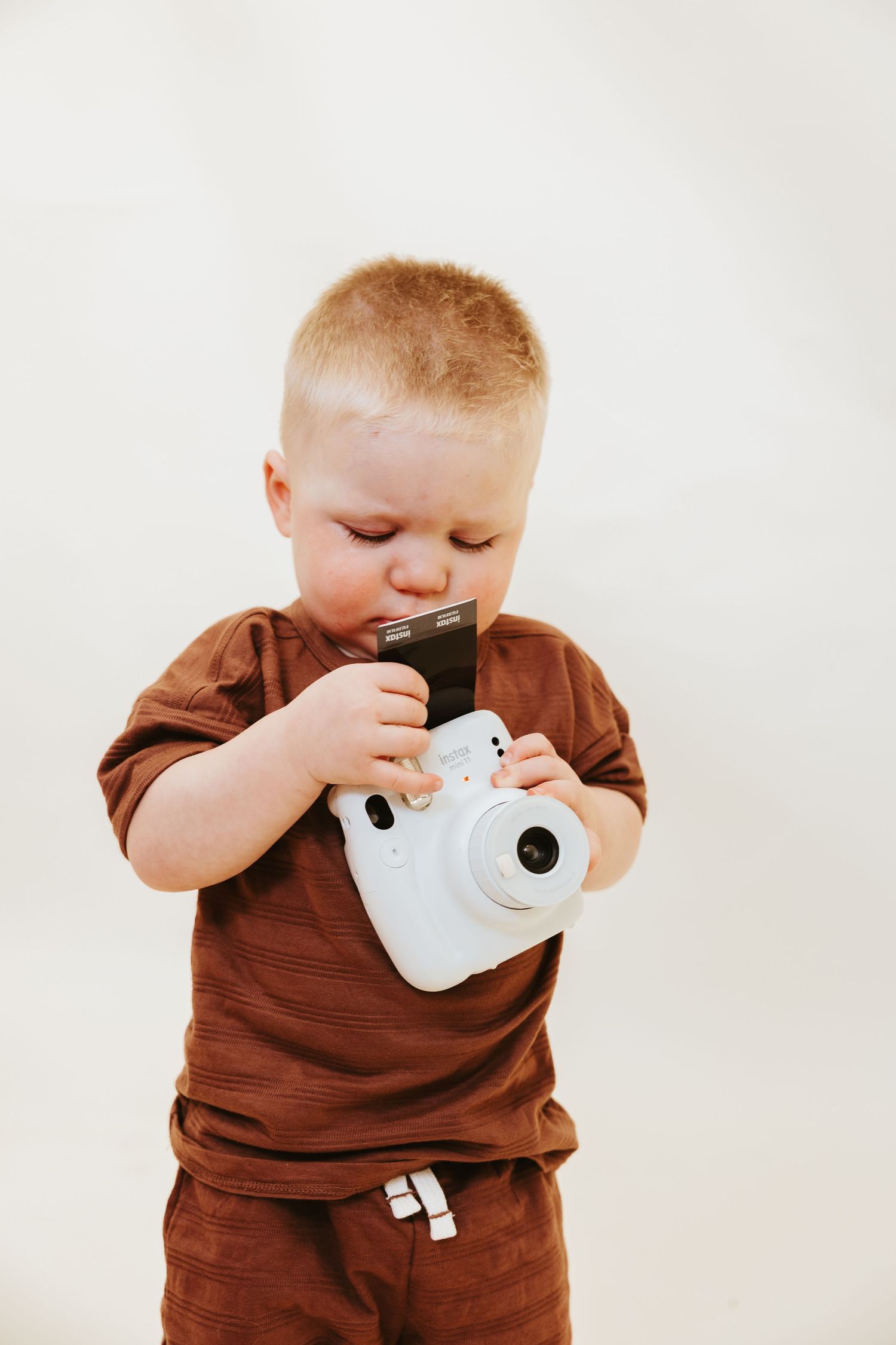 A toddler in brown clothing examines a white instant camera with curiosity.