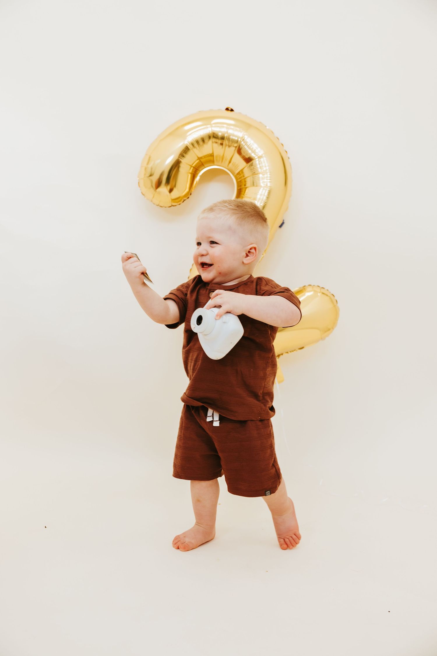 A toddler in brown clothing dances excitedly in front of a large gold number 2 balloon.