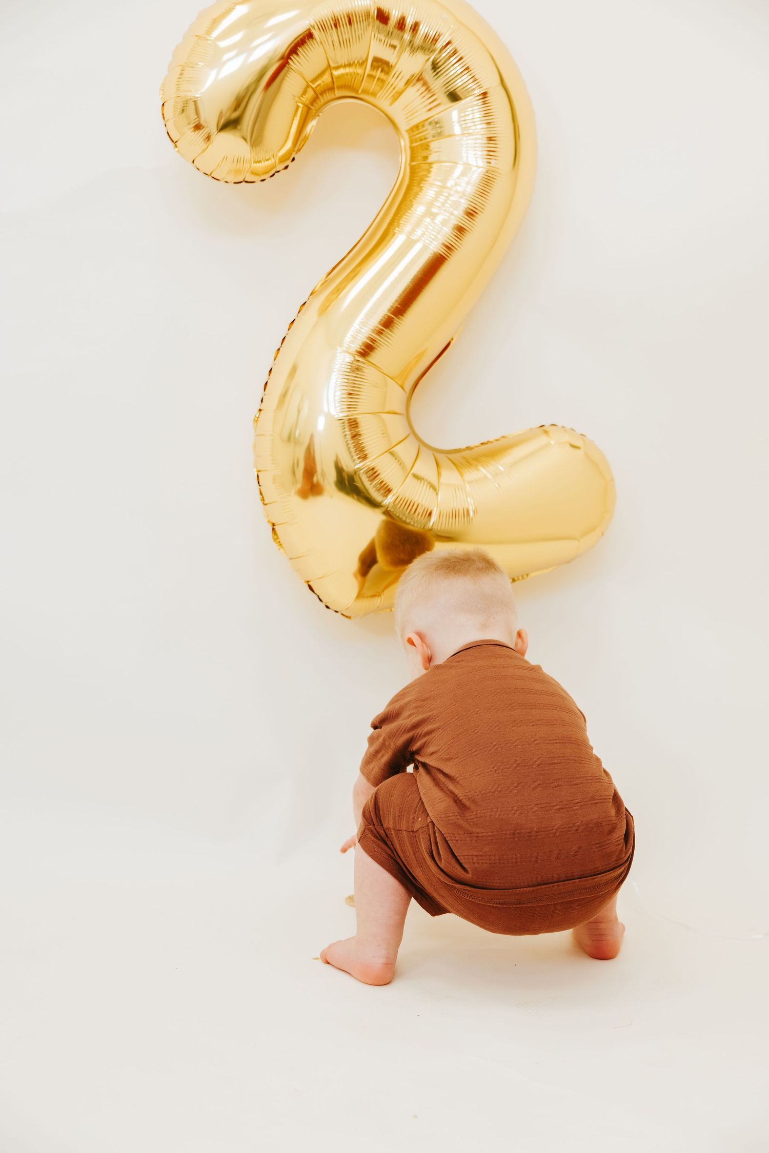 A toddler in brown clothing crouches down while looking up at a gold number 2 balloon.