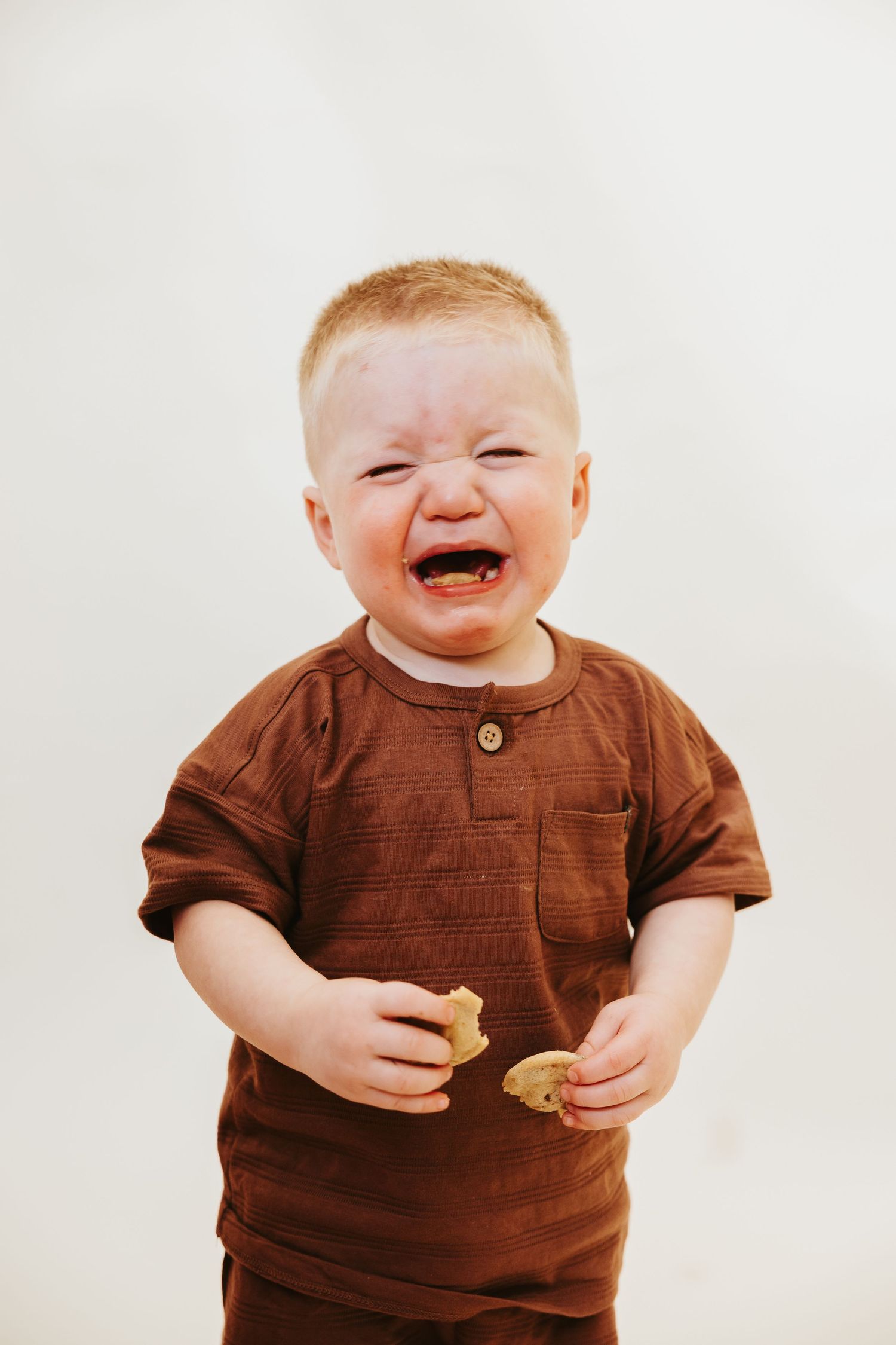 A laughing toddler in a brown outfit holds something while standing against a white background.