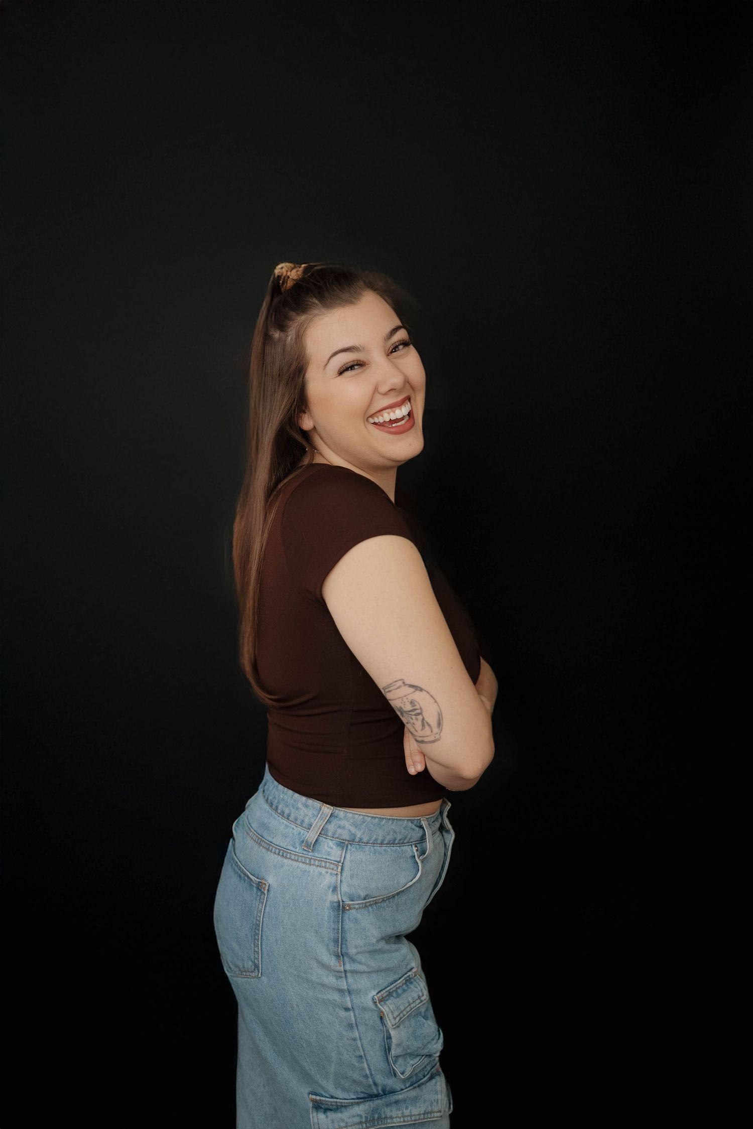 A person in a brown crop top and high-waisted jeans poses playfully against a dark background.