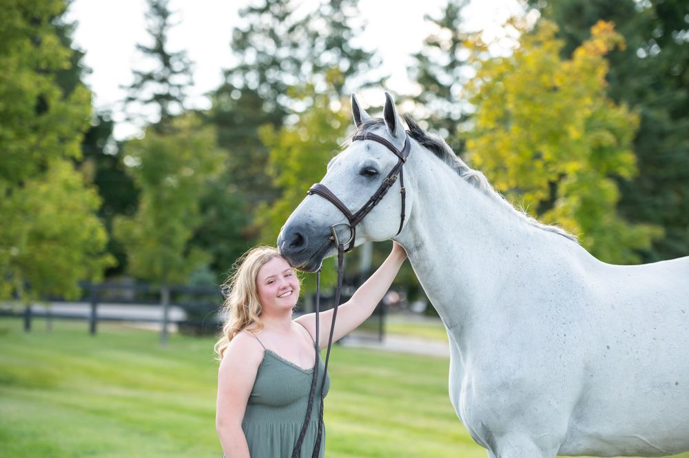 A beautiful white horse stands next to someone in a green dress on a lush summer lawn with trees in background.