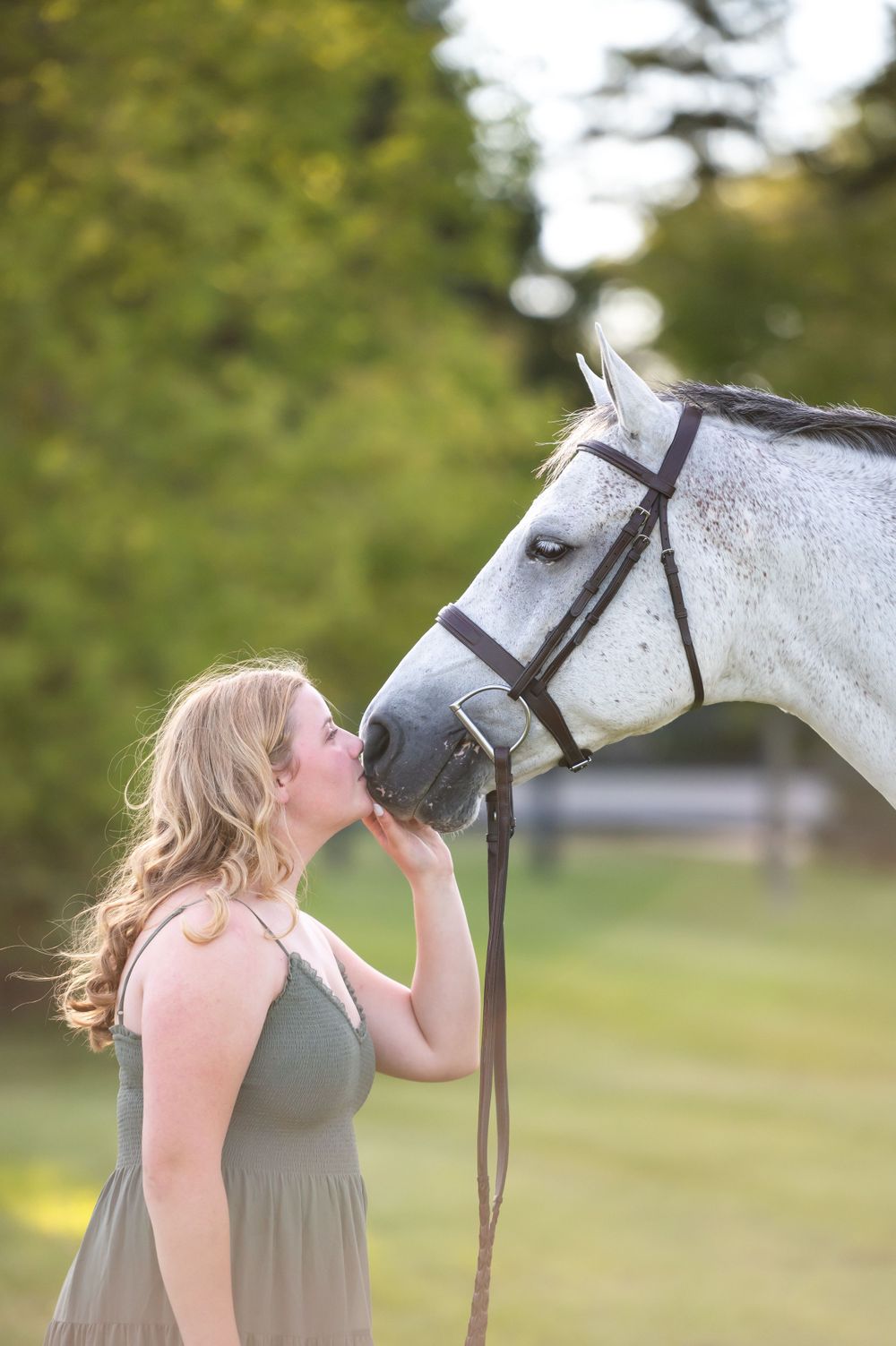 A woman in a gray dress affectionately giving a white horse a kiss on its nose in an outdoor summer setting.