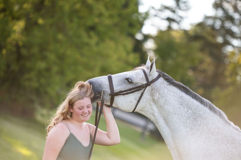 A white horse nuzzles close to a young woman outdoors in a green field during a sunny day.