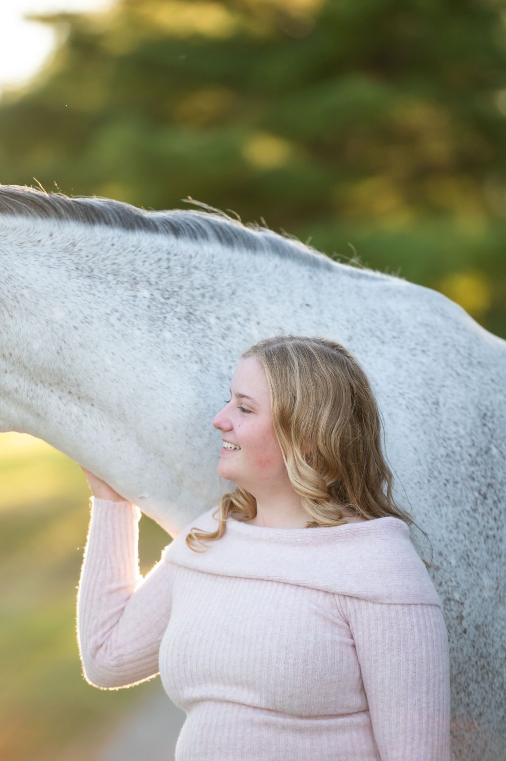 A tender moment between a white horse and a person in a pink sweater against a soft, blurred outdoor background.