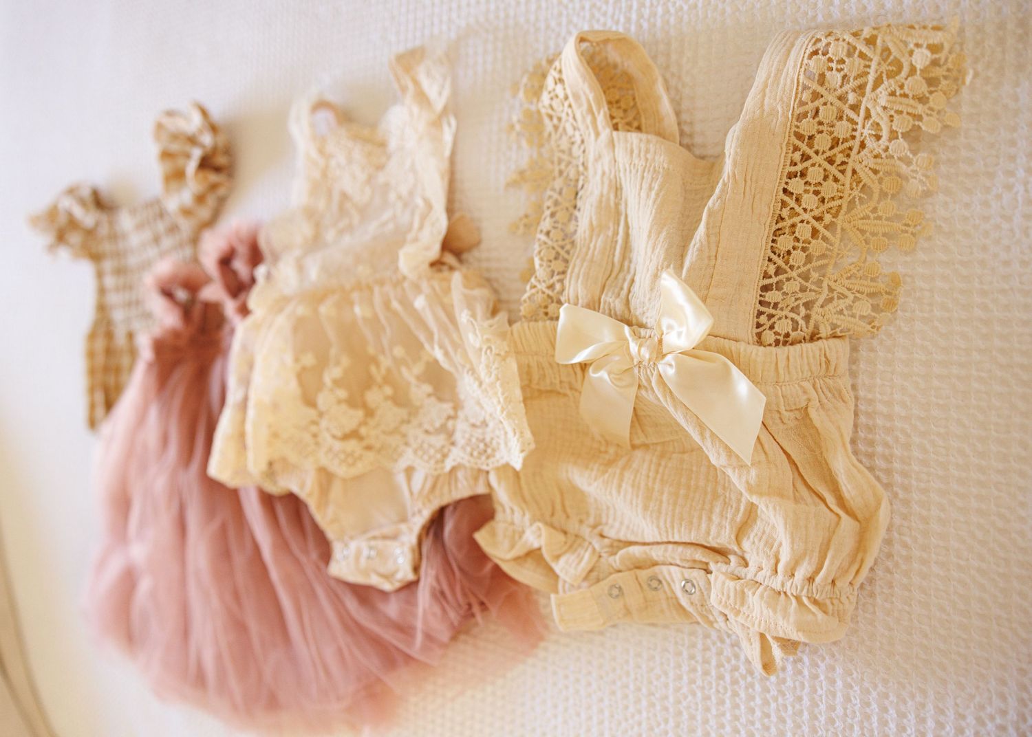 Vintage lace dresses and delicate garments hanging on a white wall in soft pastel pink and cream colors.