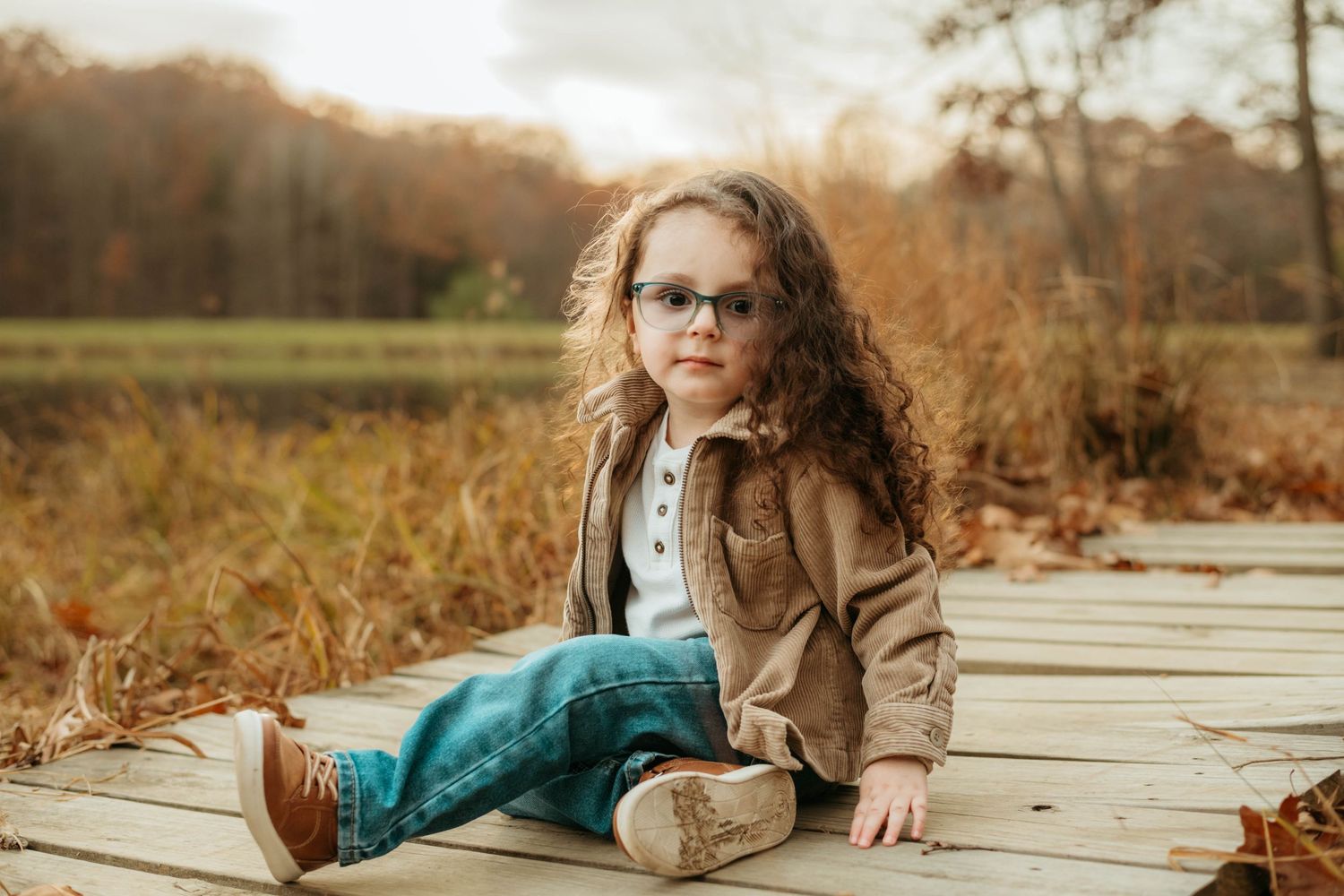 Child in brown jacket and teal pants sitting on wooden boardwalk during fall.