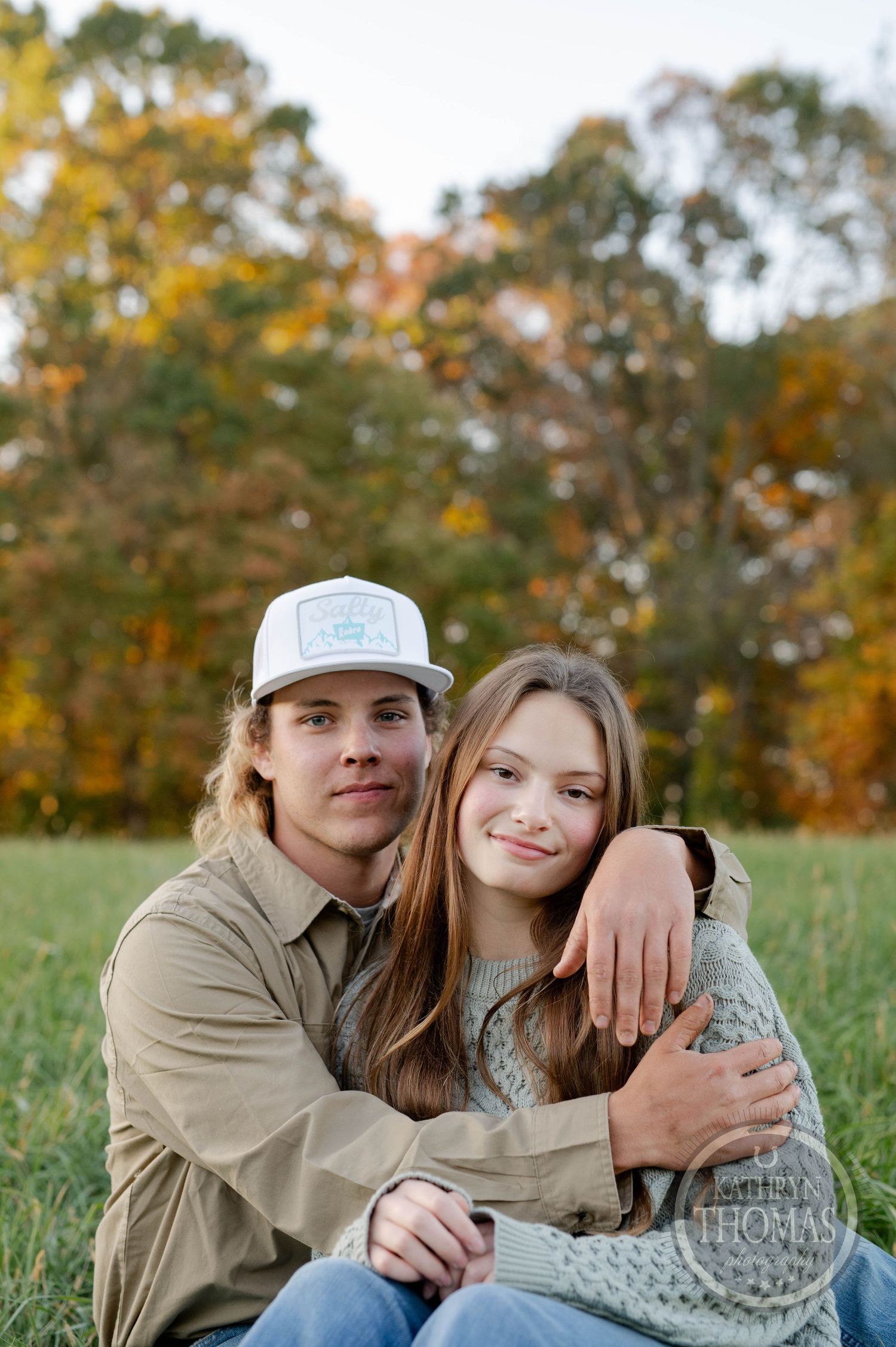 A couple embraces while sitting outdoors against a backdrop of autumn trees.