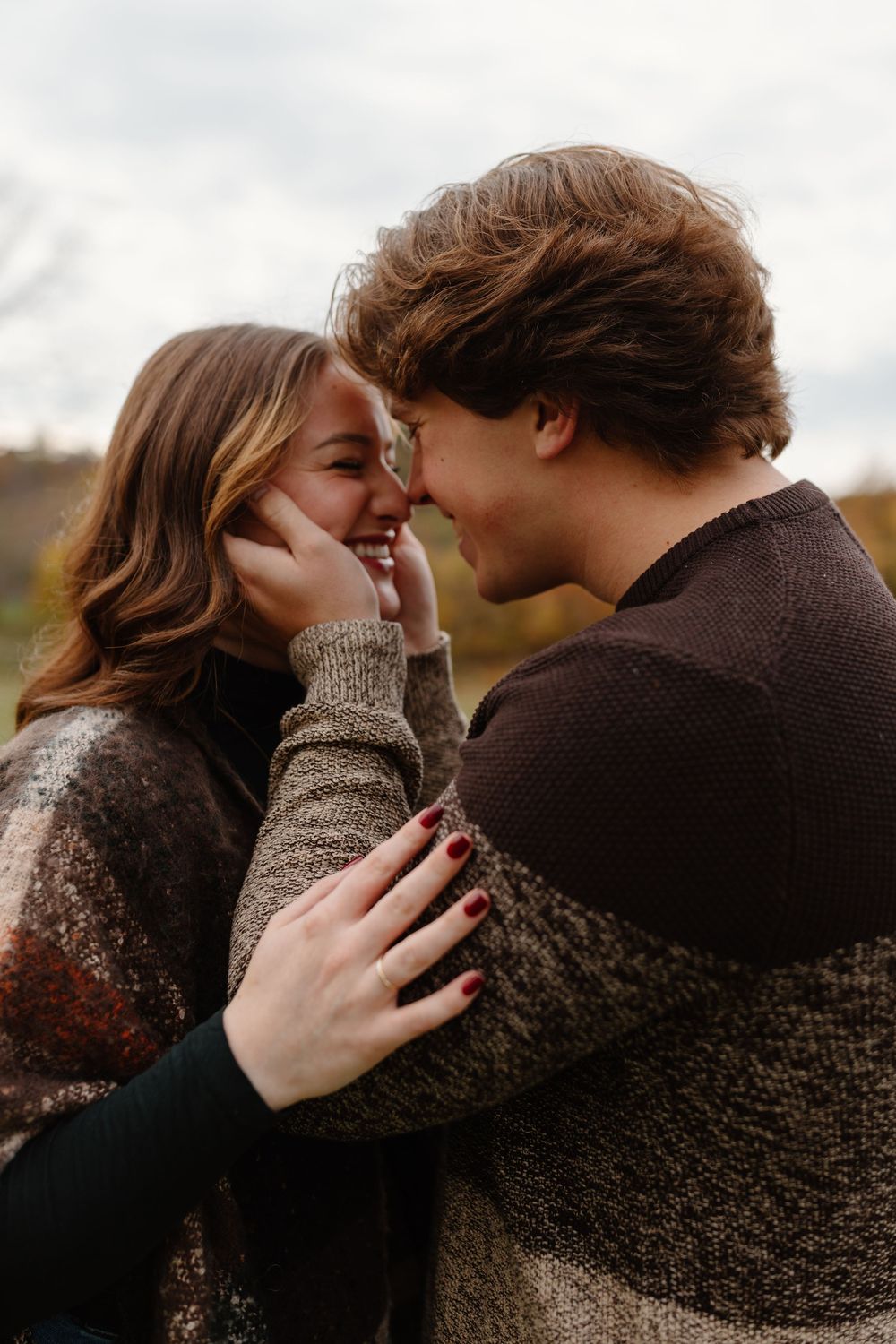 A young couple in cozy sweaters share a romantic moment outdoors, smiling and touching foreheads affectionately.
