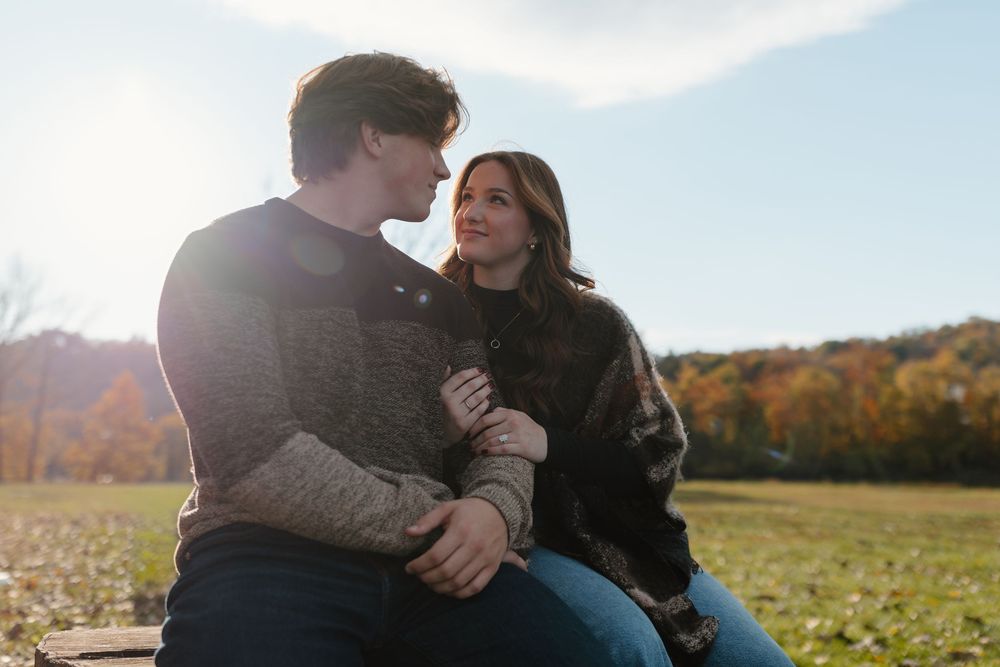 A couple sits closely together in a sunny autumn park, gazing at each other romantically.