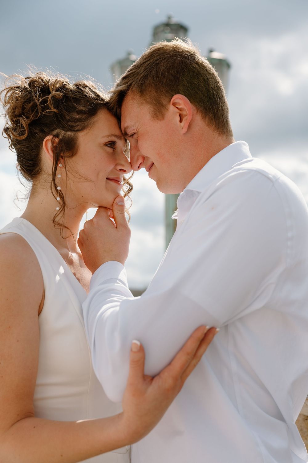 A bride and groom share a romantic forehead touch, dressed in white, with a lighthouse in the cloudy sky background.