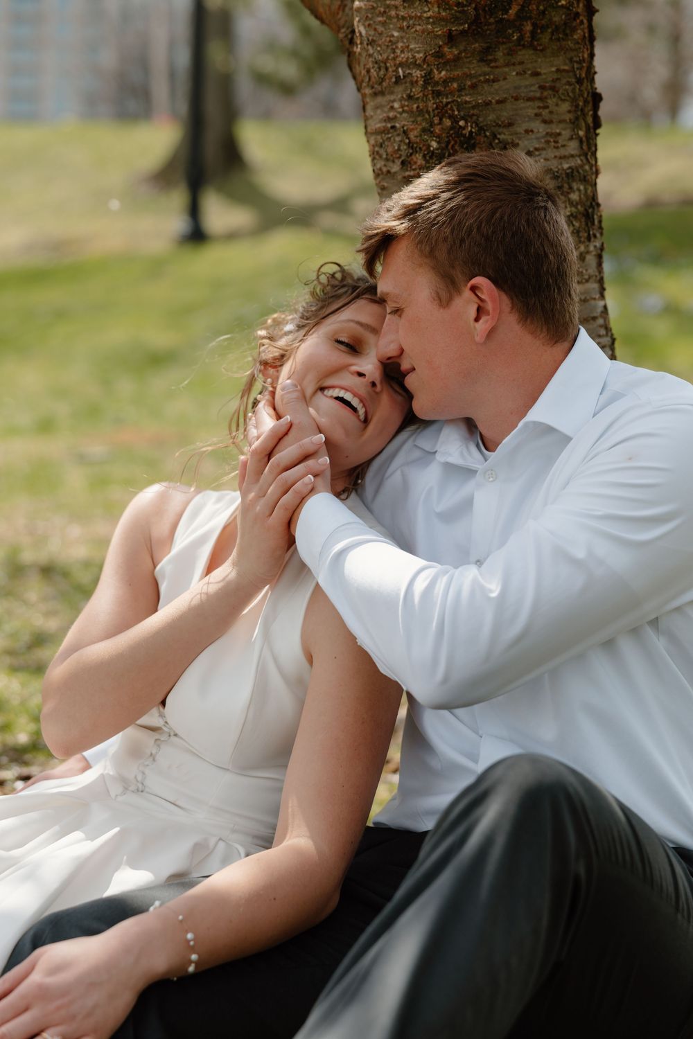 A joyful bride and groom share a tender moment seated under a tree in a sunny park setting.