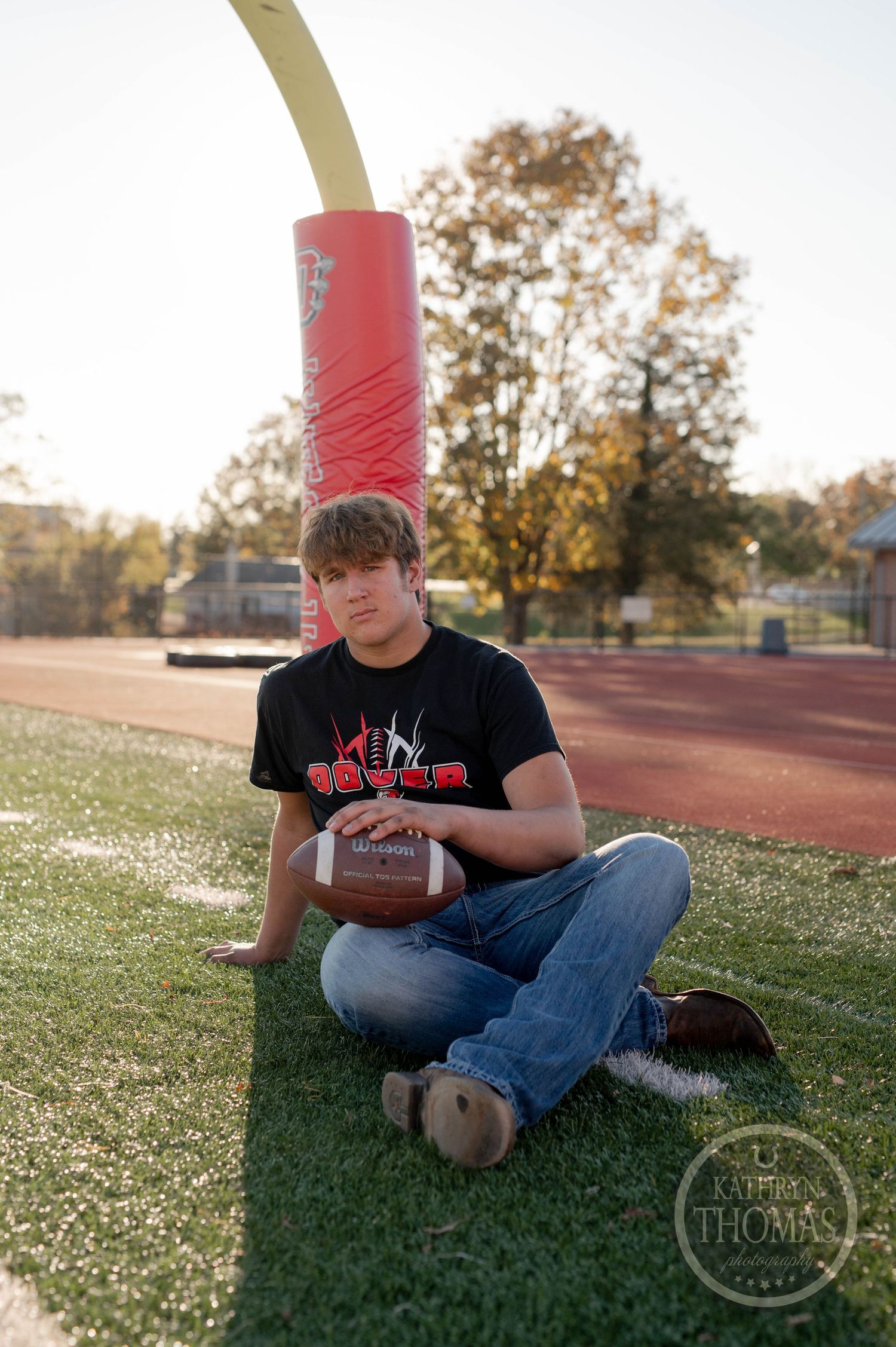Football player in black t-shirt sits on field holding football with red goalpost in background during sunset.