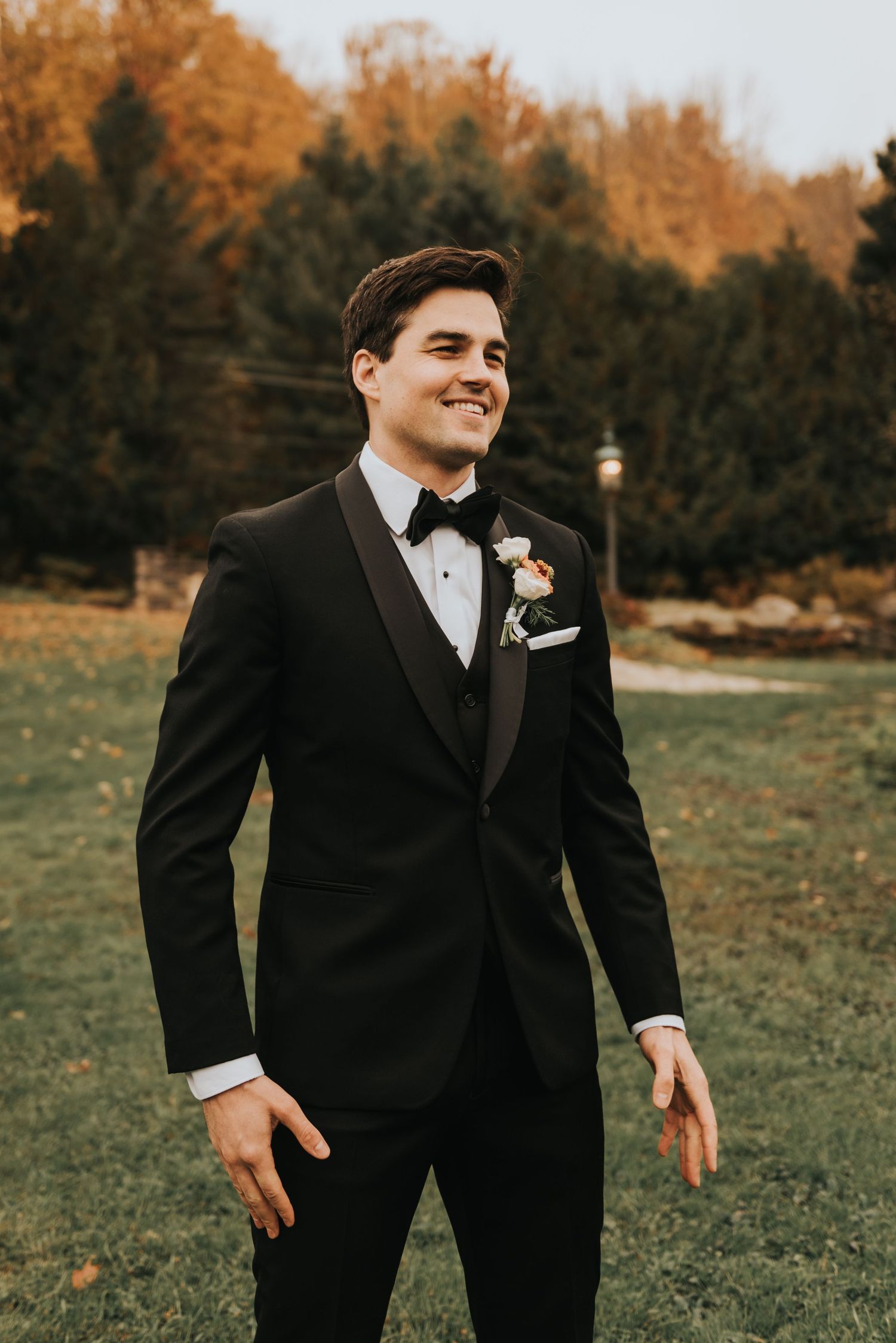An elegantly dressed person in a black tuxedo walks across a grassy field during autumn.