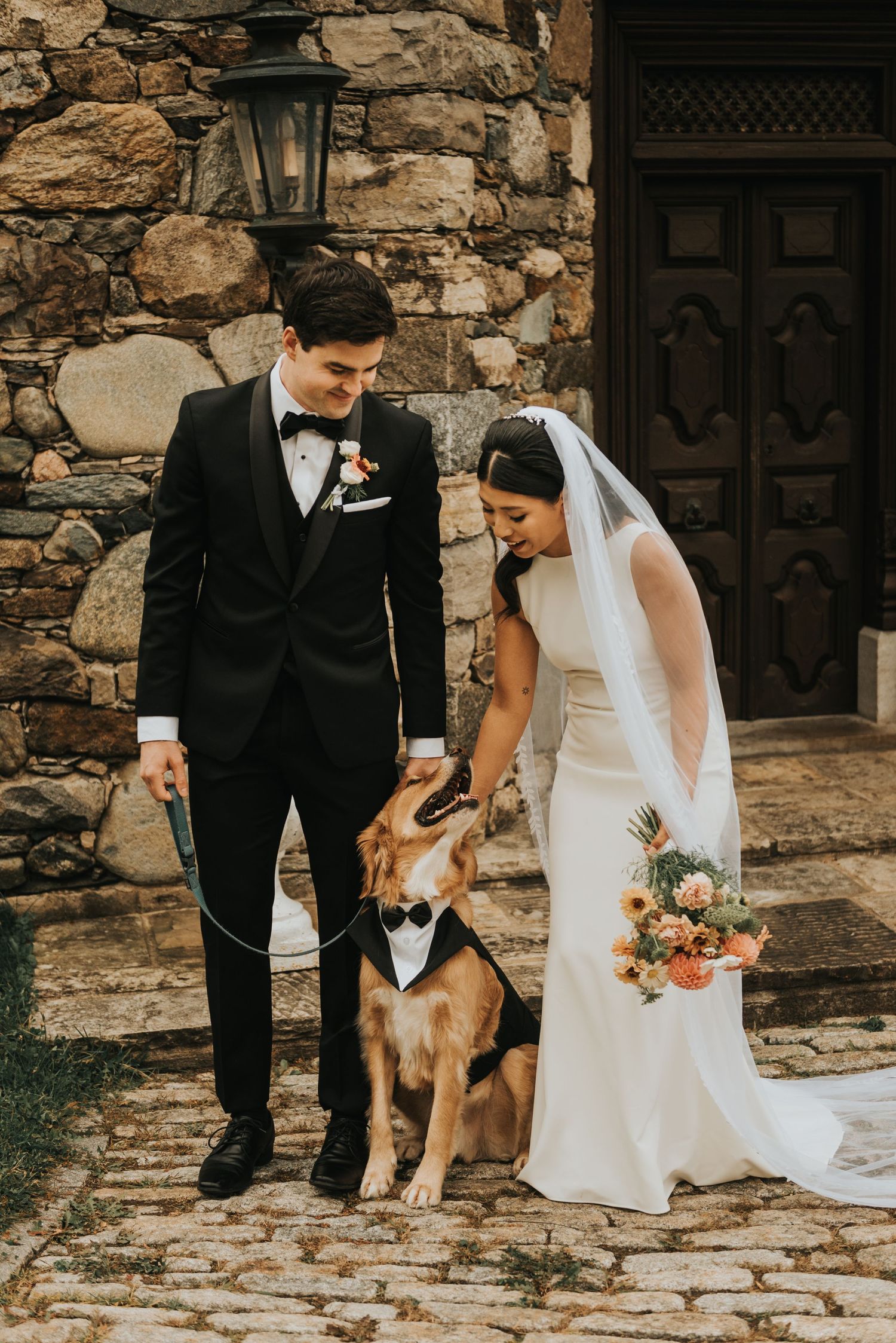 A wedding couple in formal attire poses with their dog on stone steps outside a rustic building.