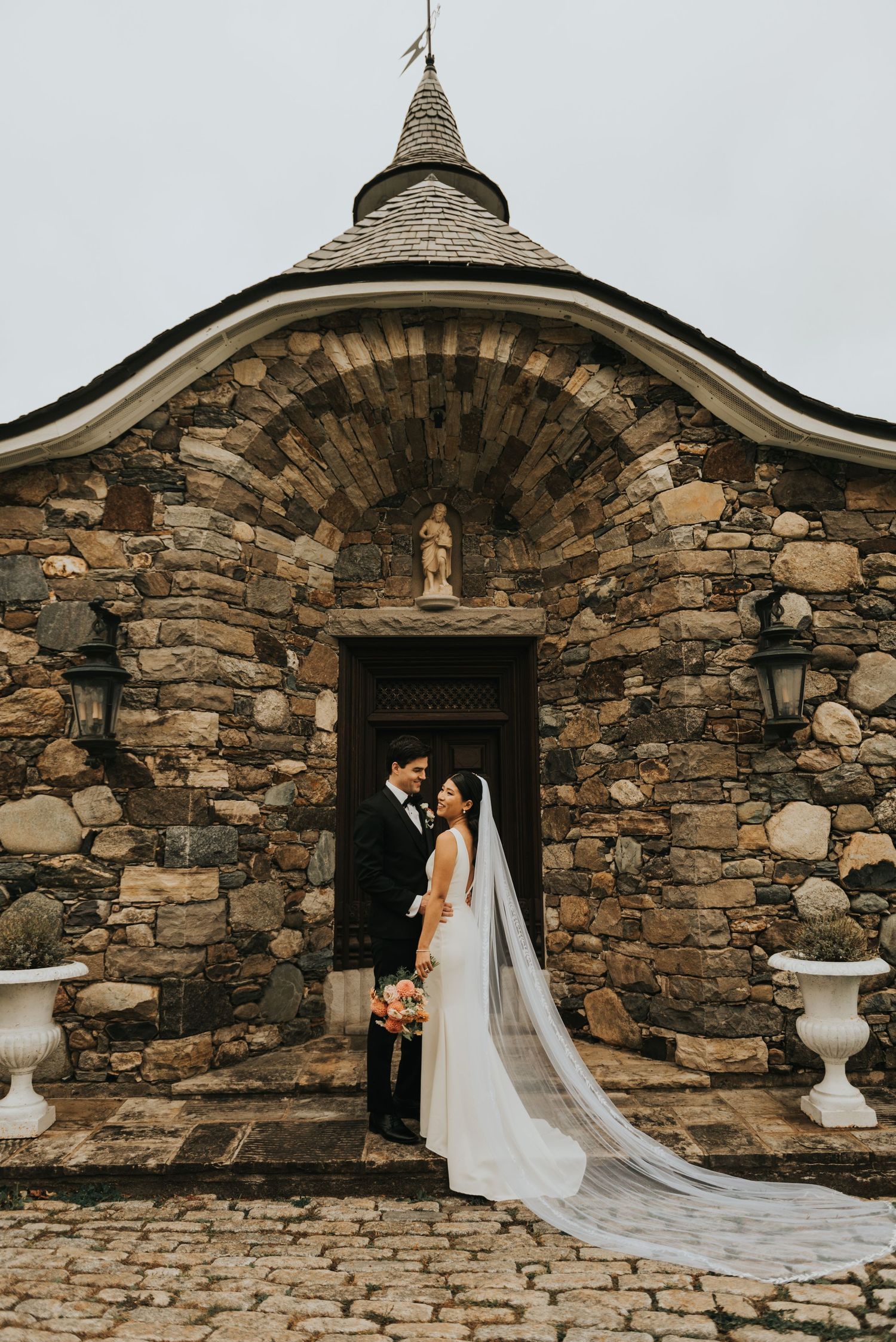 A romantic wedding couple poses in front of a rustic stone chapel doorway with a long flowing veil.