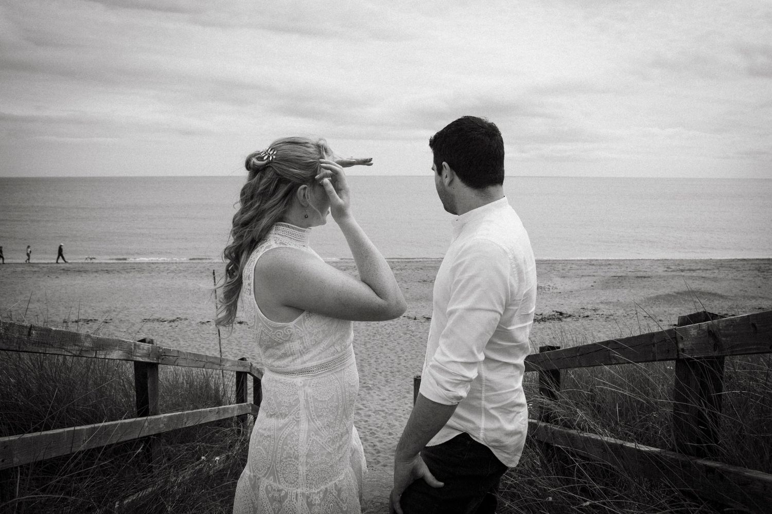 A couple stands on a coastal boardwalk looking out at the ocean in black and white photography.