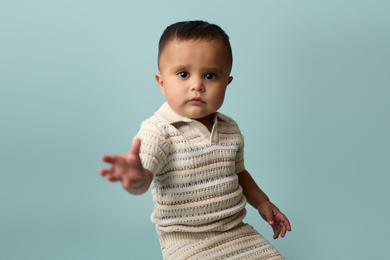 Toddler wearing cream-colored knit outfit stands against light blue background during studio portrait session.