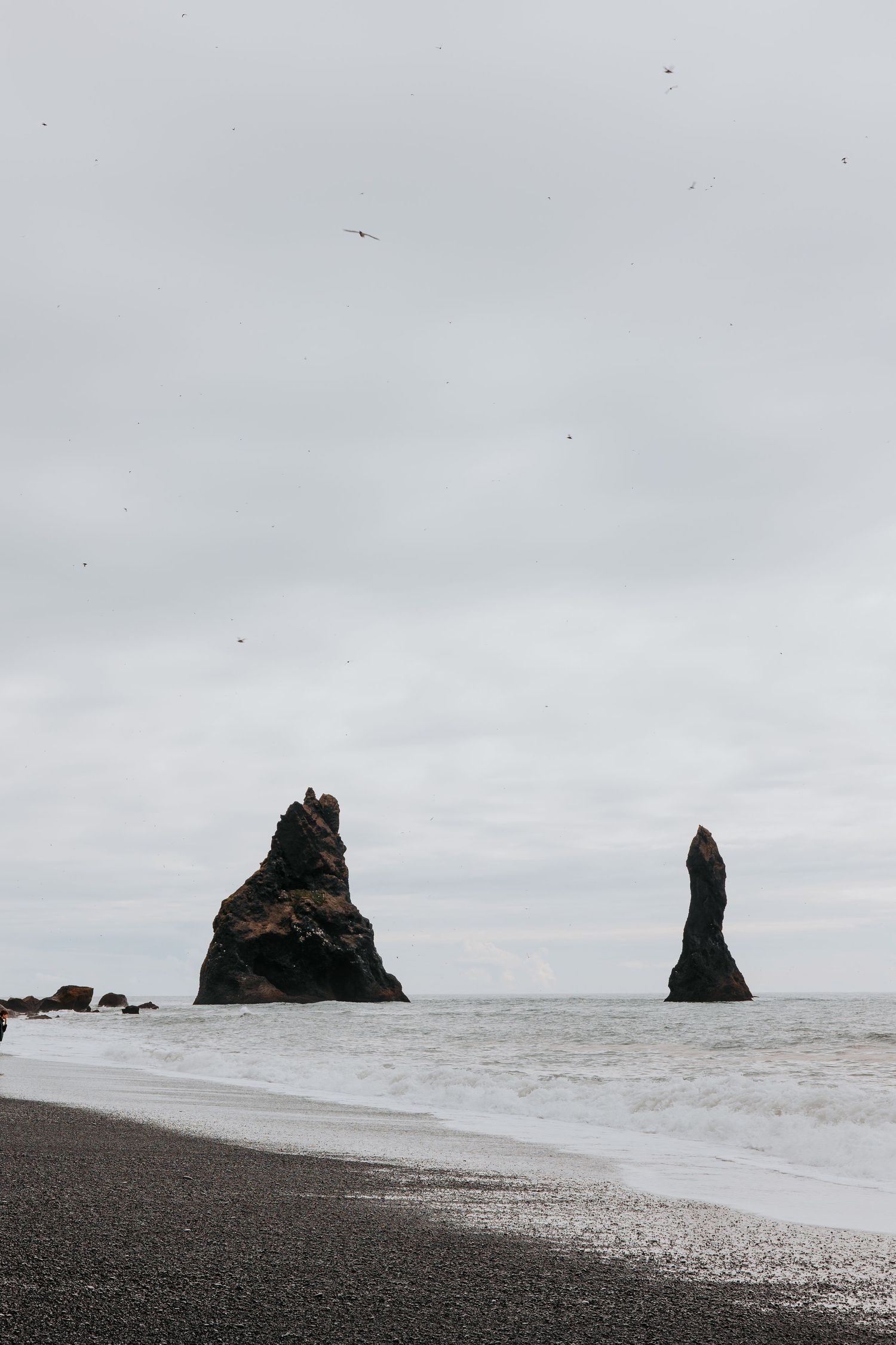 Dramatic rock formations emerge from the ocean along a remote beach under grey skies.
