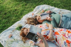 Portrait of two women holding books up to their noses while laying on a blanket covered in flowers.