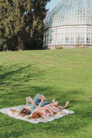 Candid photo of two women relaxing on a blanket in front of a botanical conservatory while reading.