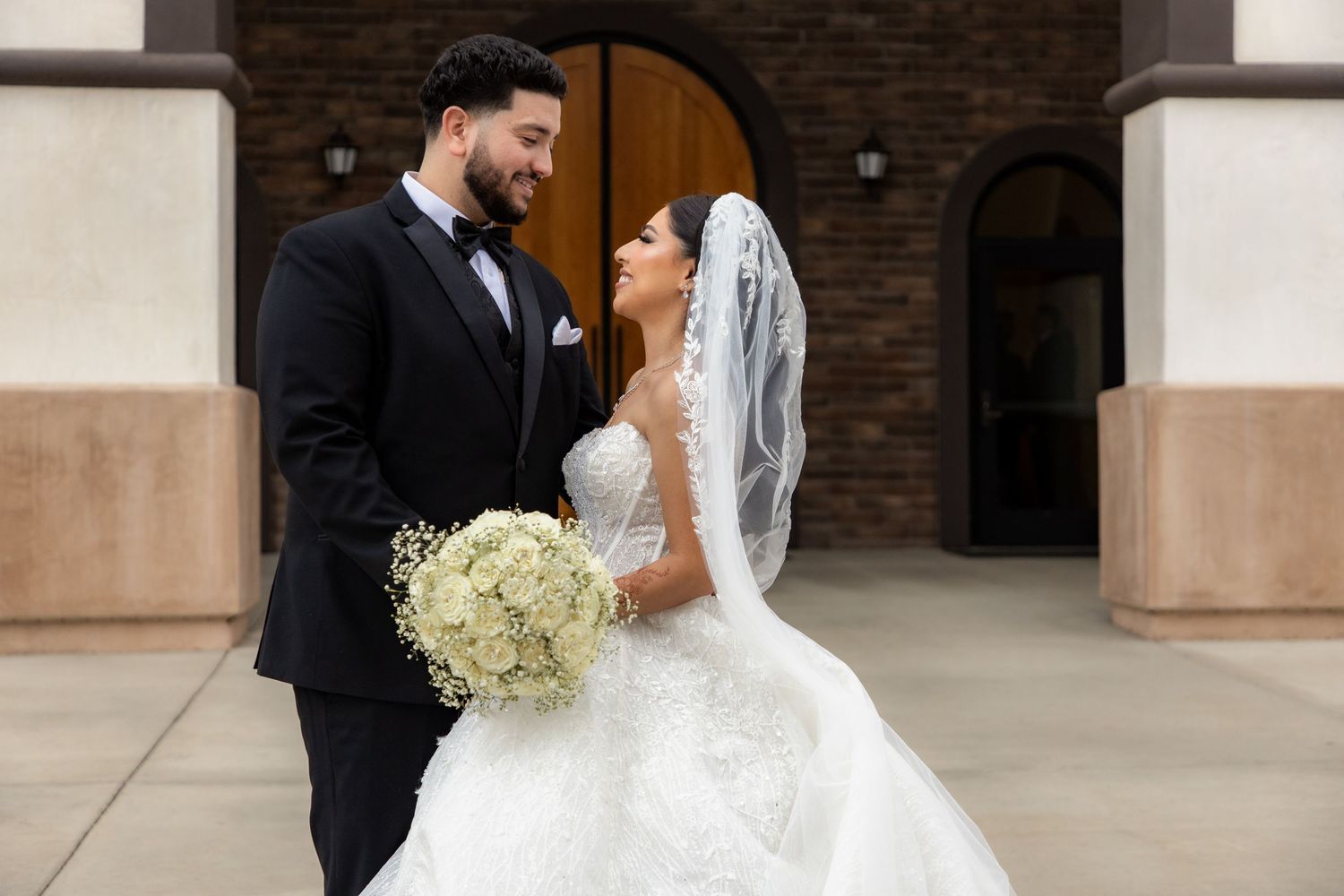 Elegant bride in lace ball gown and groom in black tuxedo share a loving gaze outside a church.