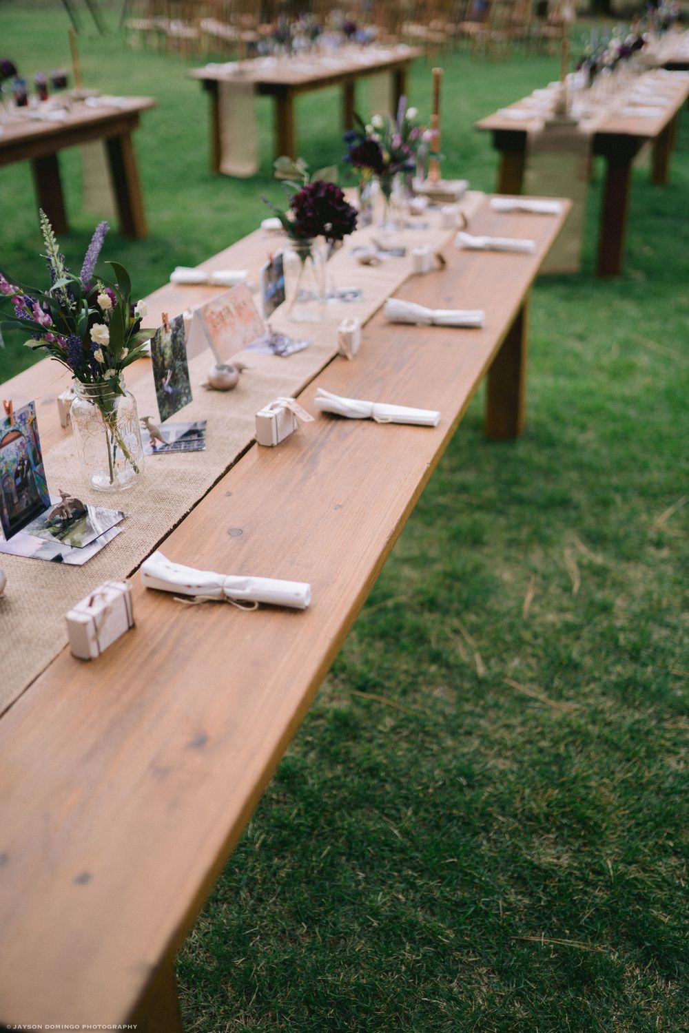 Rustic wooden tables set for outdoor dining with simple floral centerpieces and place settings on a grass lawn.