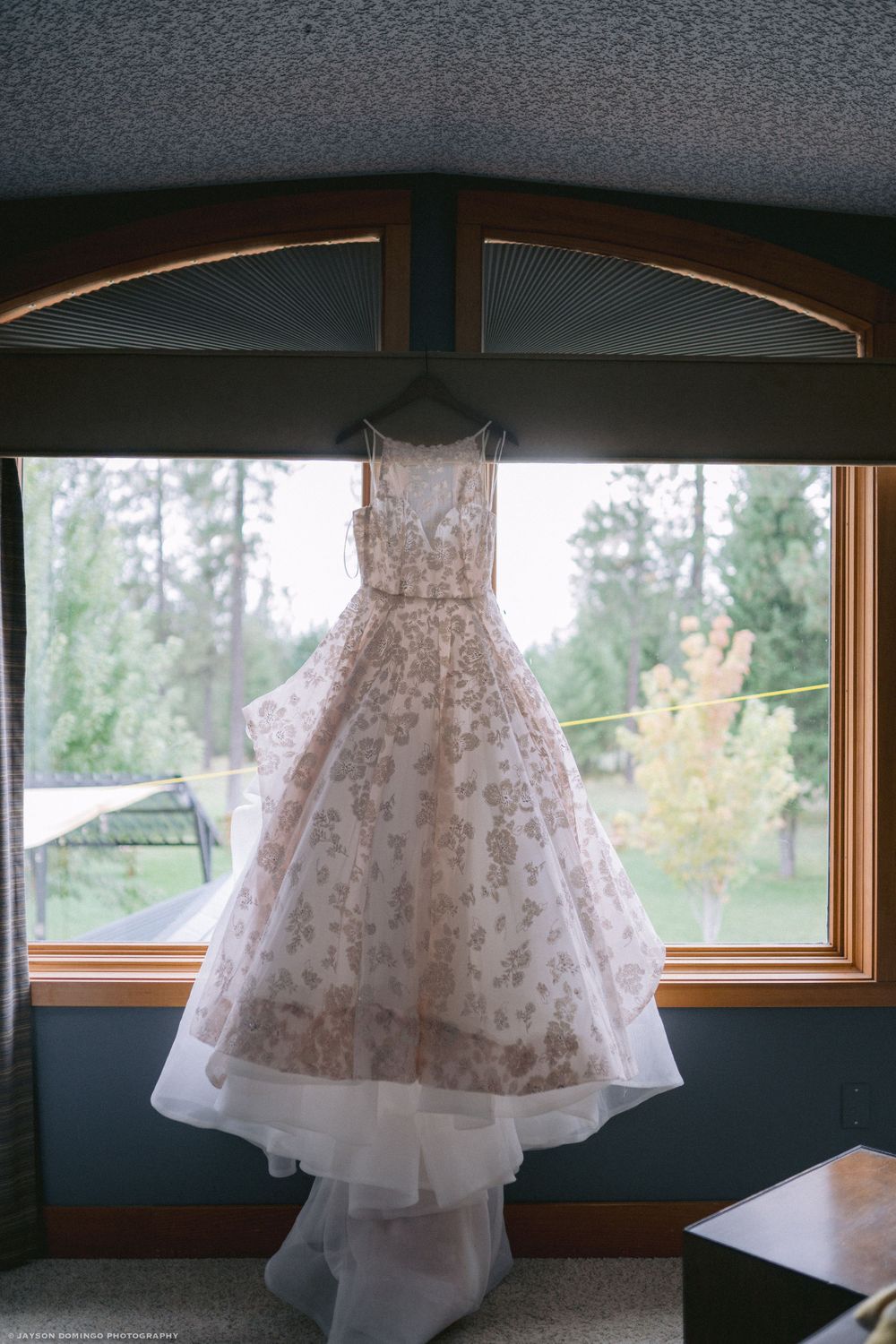 A delicate wedding dress with lace details hangs in front of a window with forest views.