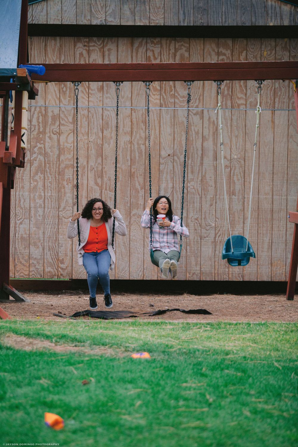Two people enjoying swings mounted on a wooden barn wall with a grassy lawn in the foreground.