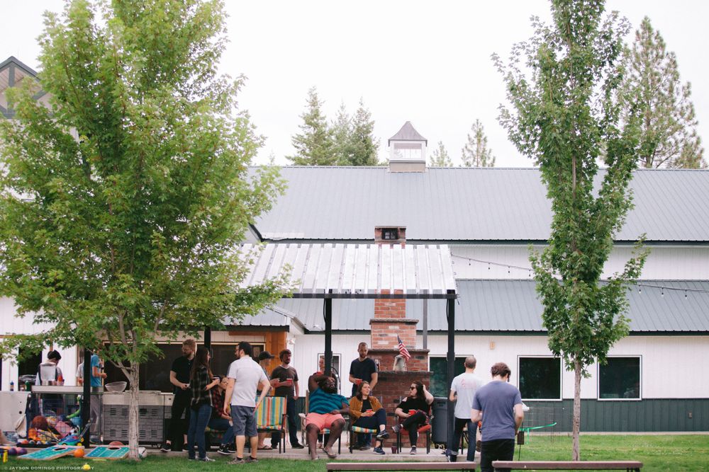 People gather outside a white barn building with tall green trees at a rustic outdoor venue.