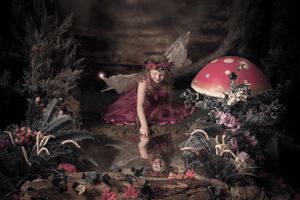 A fairy in a pink dress leans over to touch her reflection in a magical woodland pond near a red toadstool.