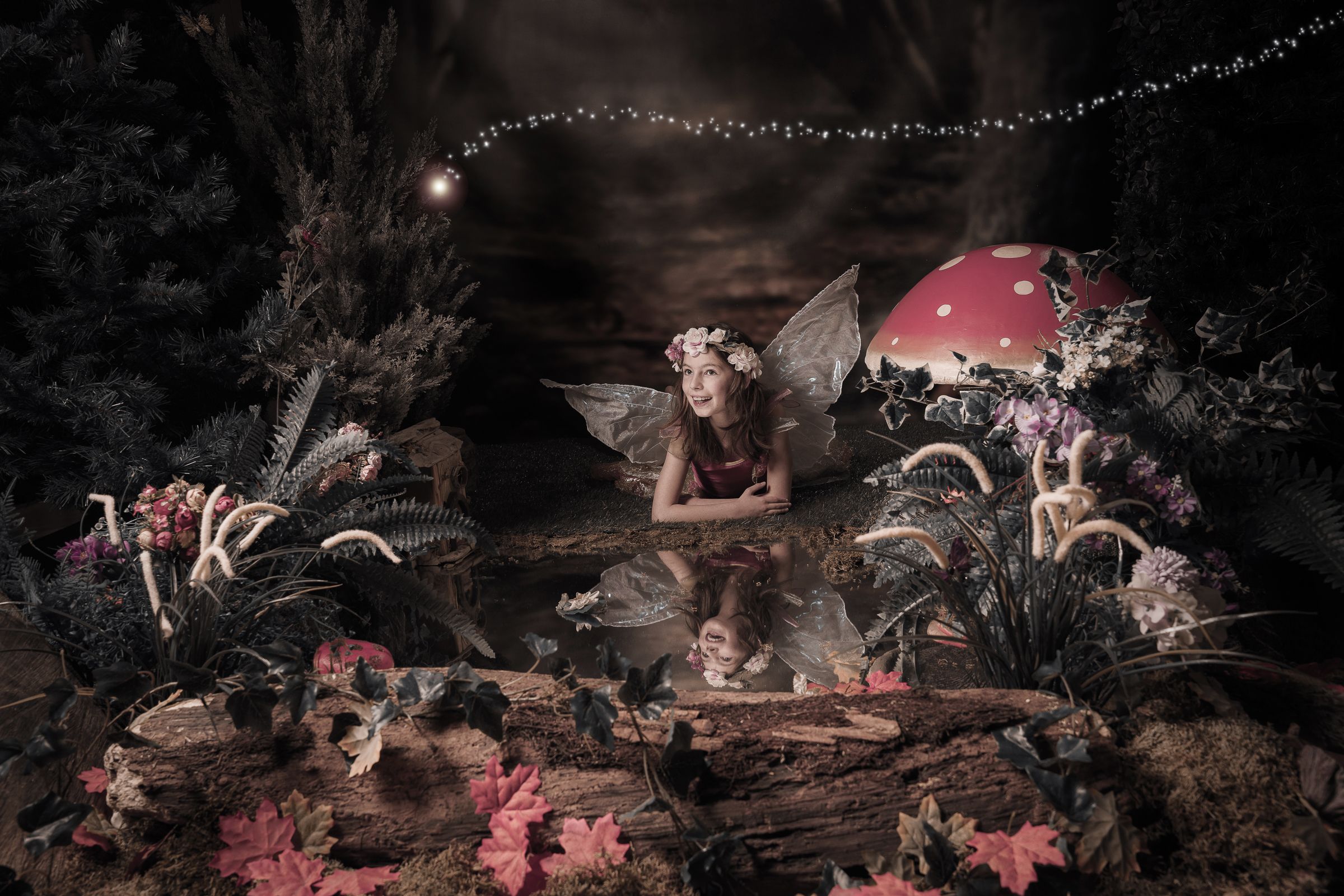 A fairy sits beneath twinkling lights in a dark woodland setting with a red mushroom and glowing flowers.