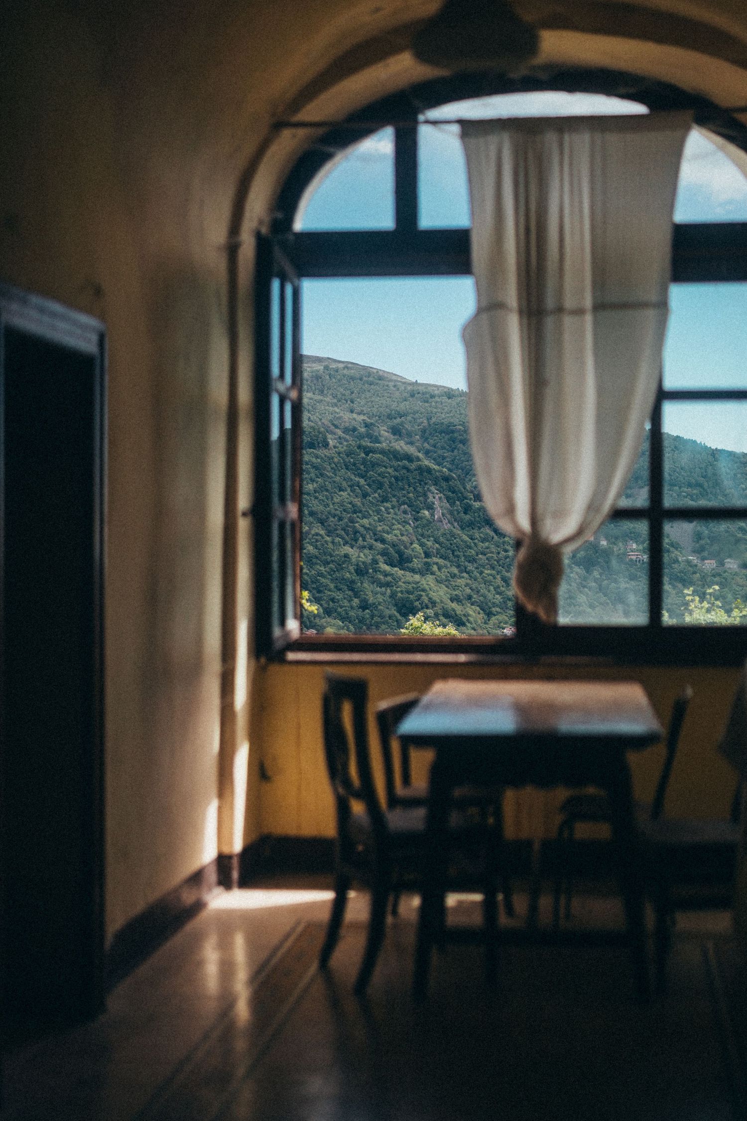 Arched window with white curtain billowing over rustic table revealing lush mountain view beyond.
