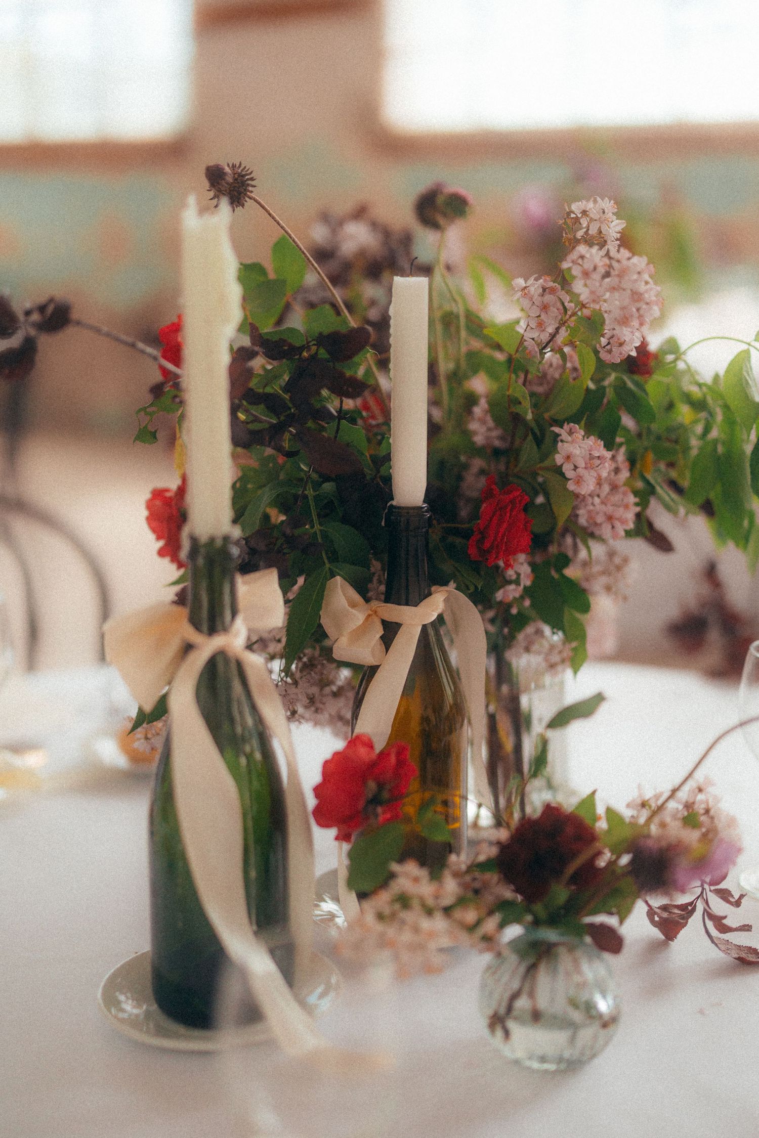 Elegant wedding table centerpiece with white candles in green bottles tied with ribbon, surrounded by colorful blooms.