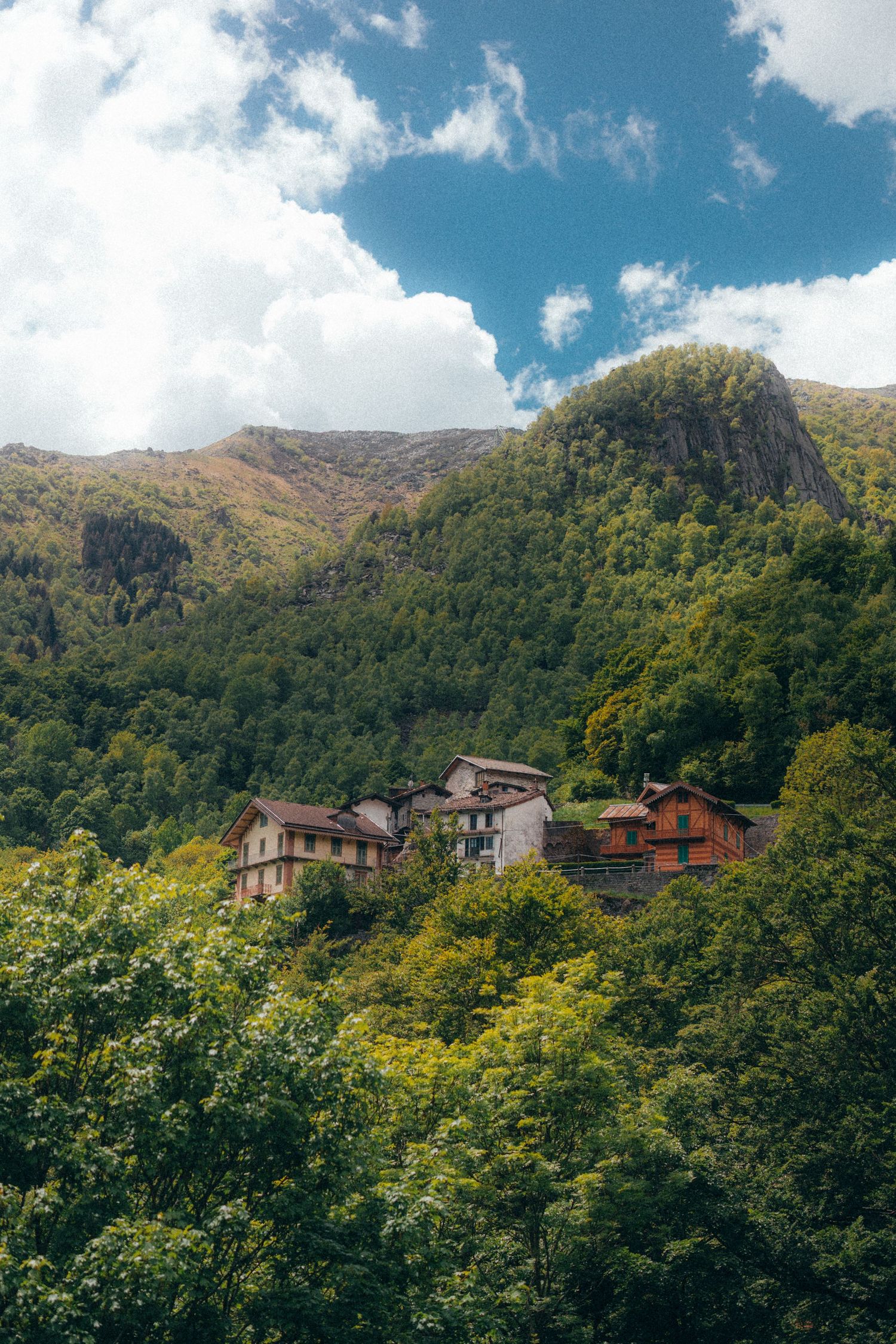 Lush green mountain village with rustic houses nestled among trees under a blue sky with white clouds.