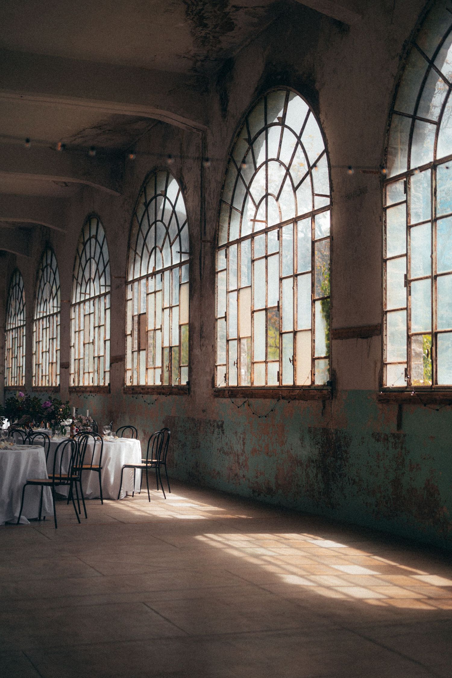 Rustic industrial wedding venue with tall arched windows, peeling walls, and elegant white-draped reception tables.