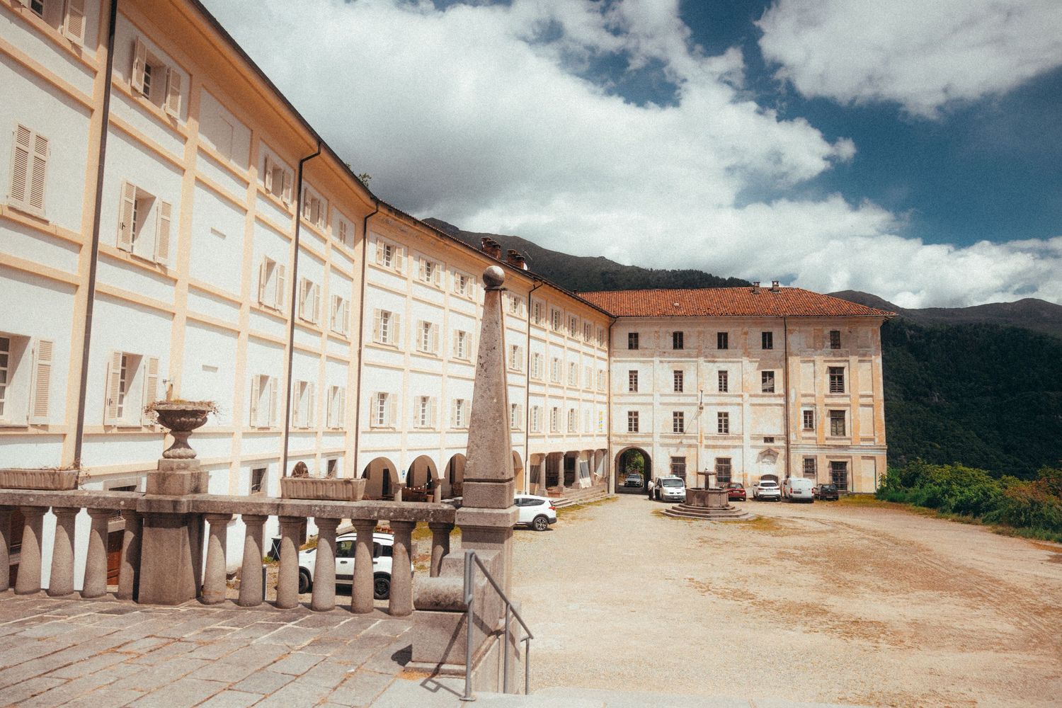 Grand historic monastery courtyard with white baroque facade, arched colonnade, and mountain backdrop.