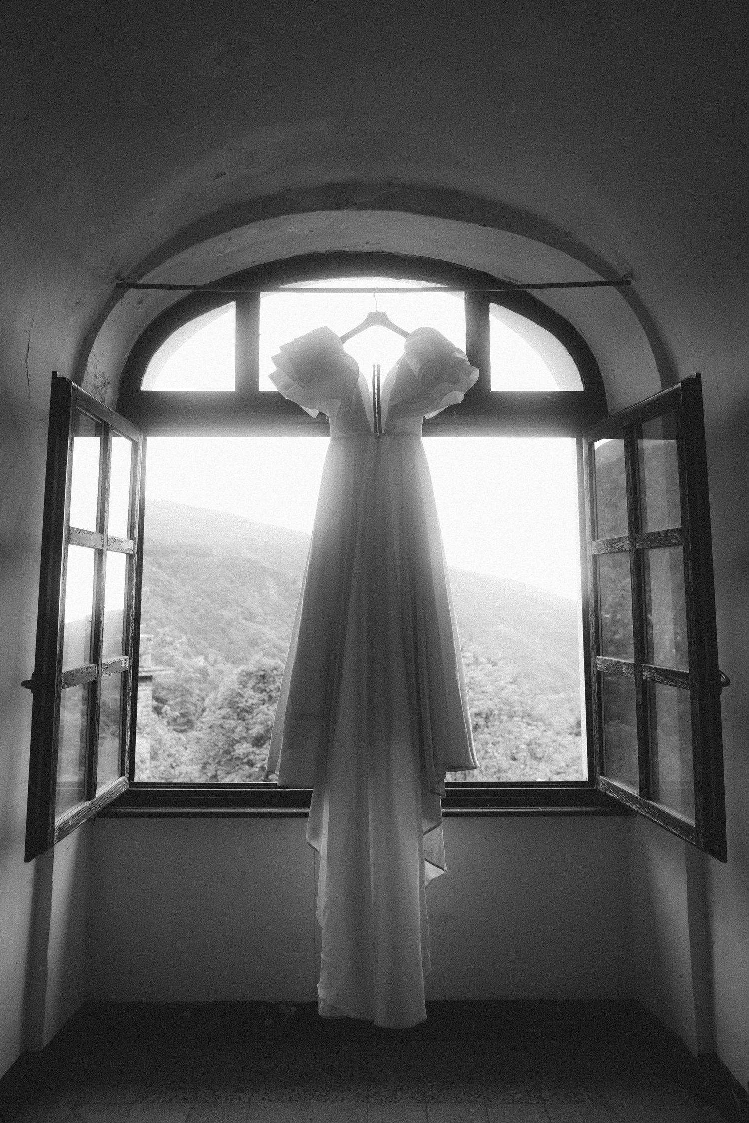 White wedding dress hanging in arched window with mountain forest view in black and white photo.